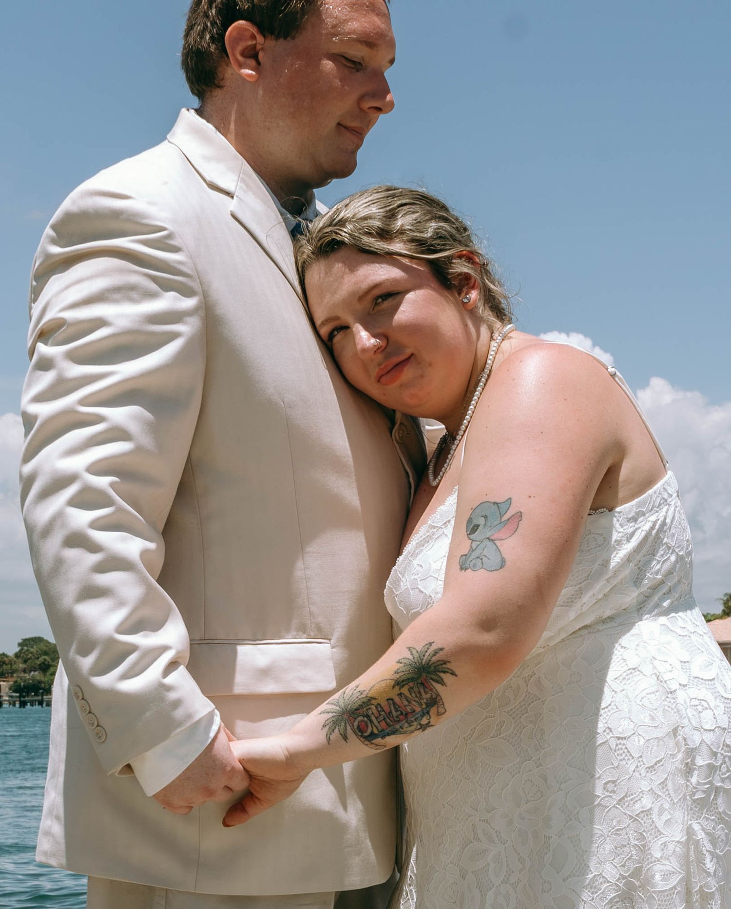 Bride with tattoos in white dress embraces groom in cream suit near waterfront on wedding day.