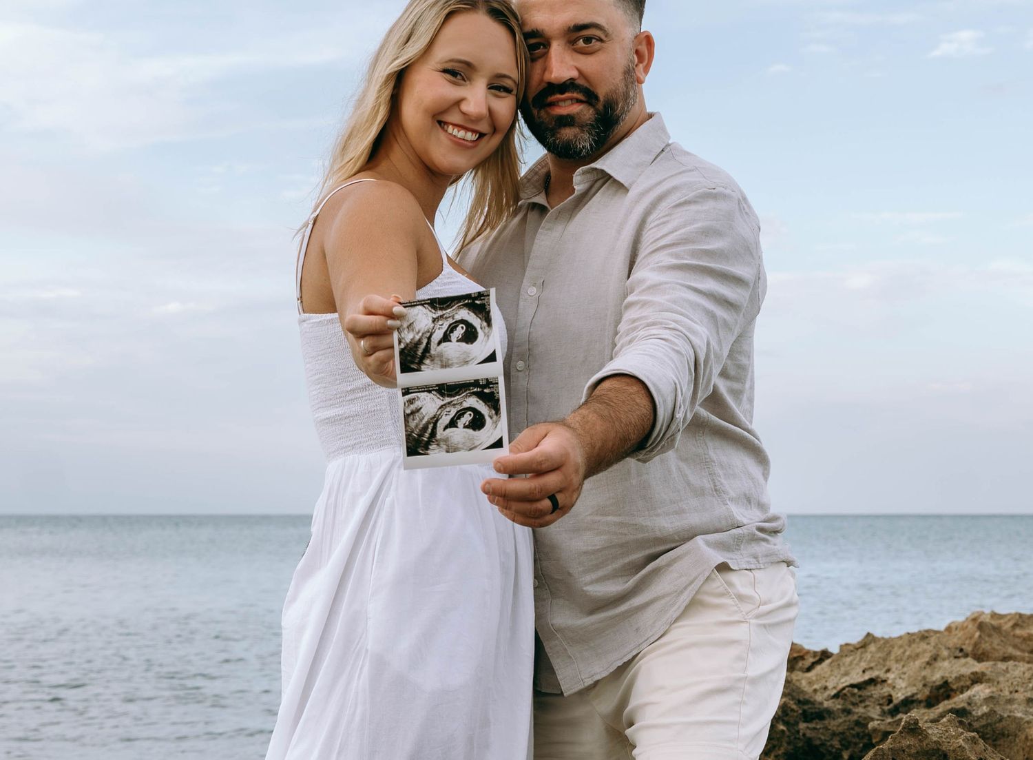 Happy couple holding ultrasound photos on rocky beach, announcing pregnancy with ocean backdrop.