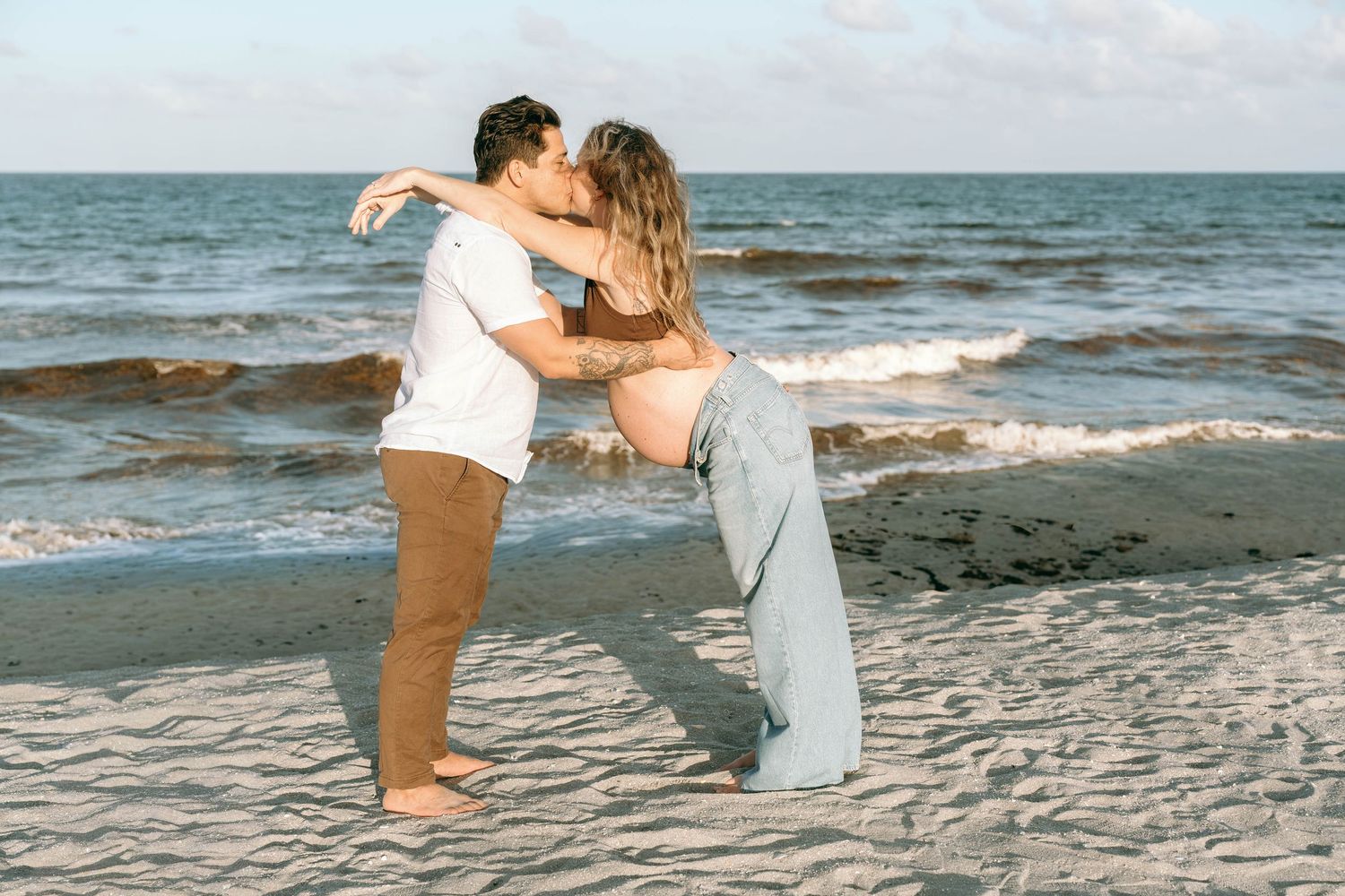 Couple kissing on a sandy beach with ocean waves, woman showing pregnant belly in blue jeans.