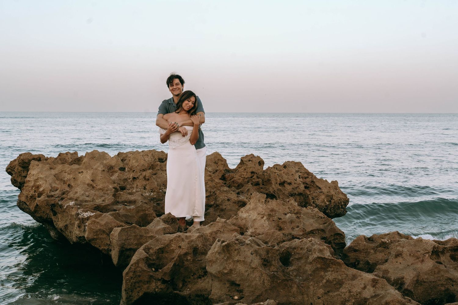 Couple embracing on rocky shoreline at sunset with calm ocean waves in the background.