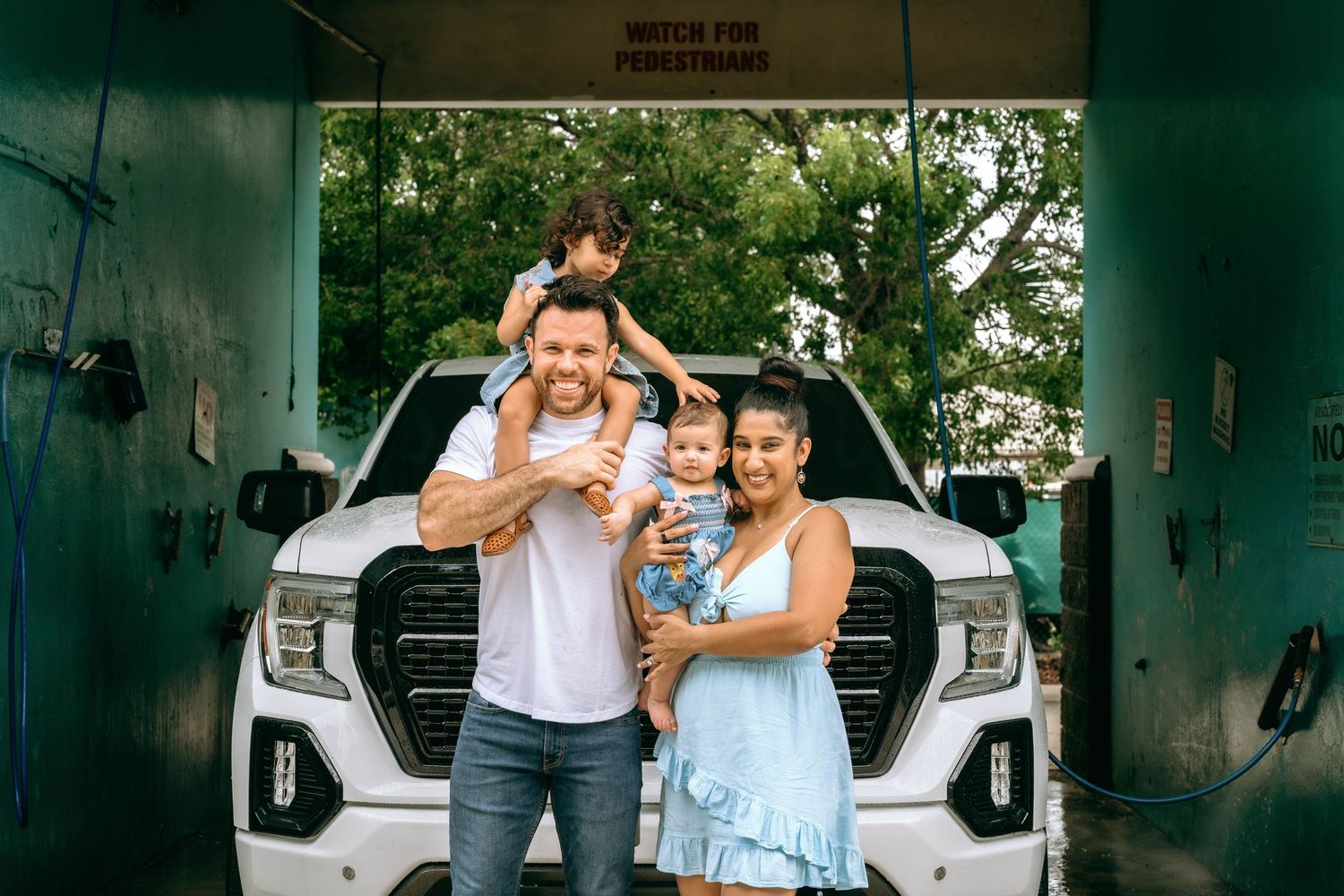 Family of four posing in front of white GMC truck at car wash, parents holding young children, smiling outdoors.