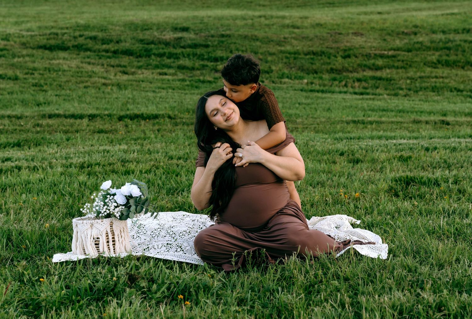 Pregnant woman sitting on white blanket in green field, embraced from behind by partner, with flower bouquet nearby.
