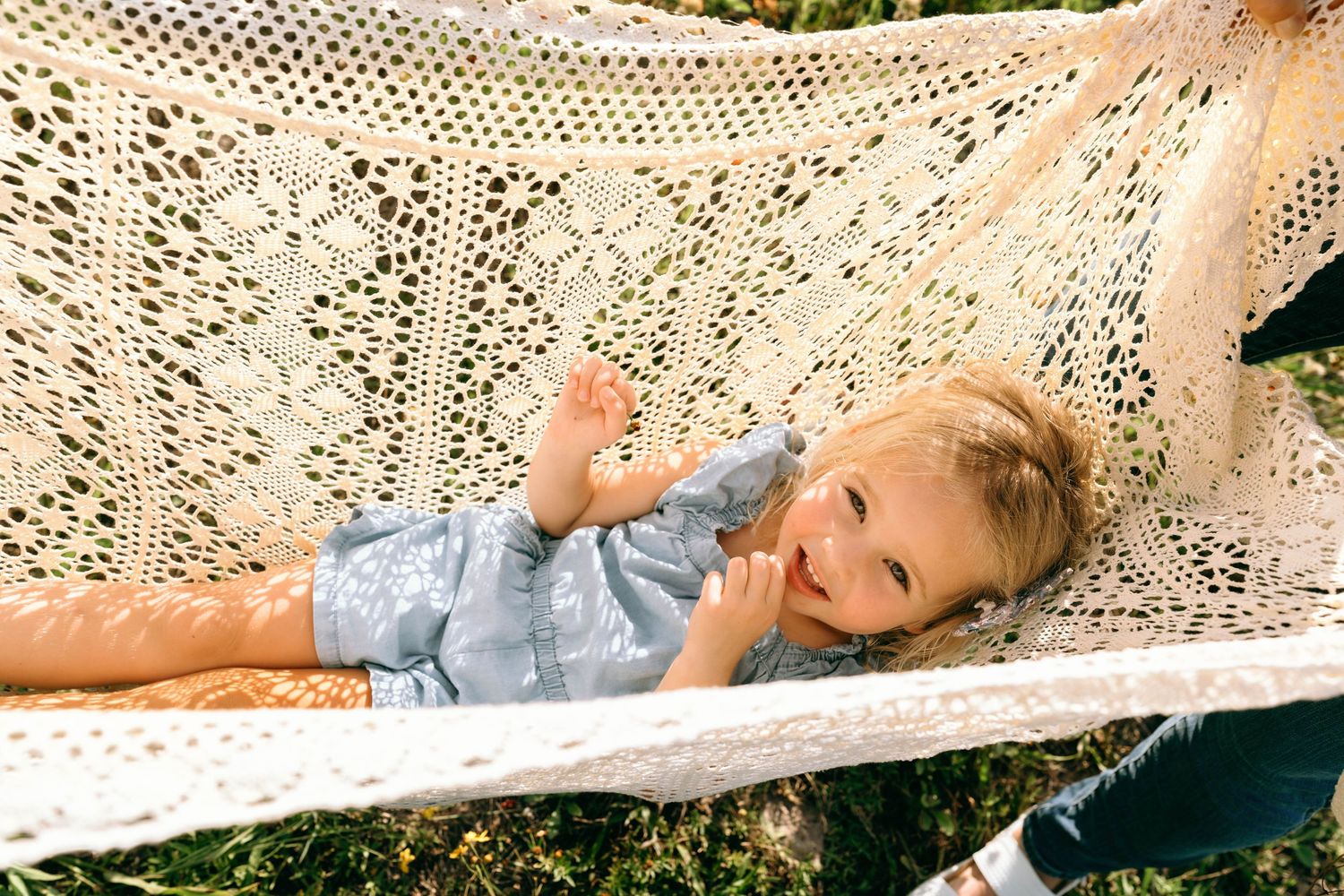 A happy toddler with blonde hair relaxes in a cream crochet hammock outdoors, smiling and looking up.