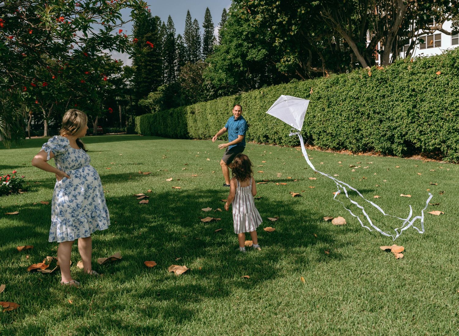 Family flying a white kite on lush green lawn surrounded by trees and hedges on a sunny day.