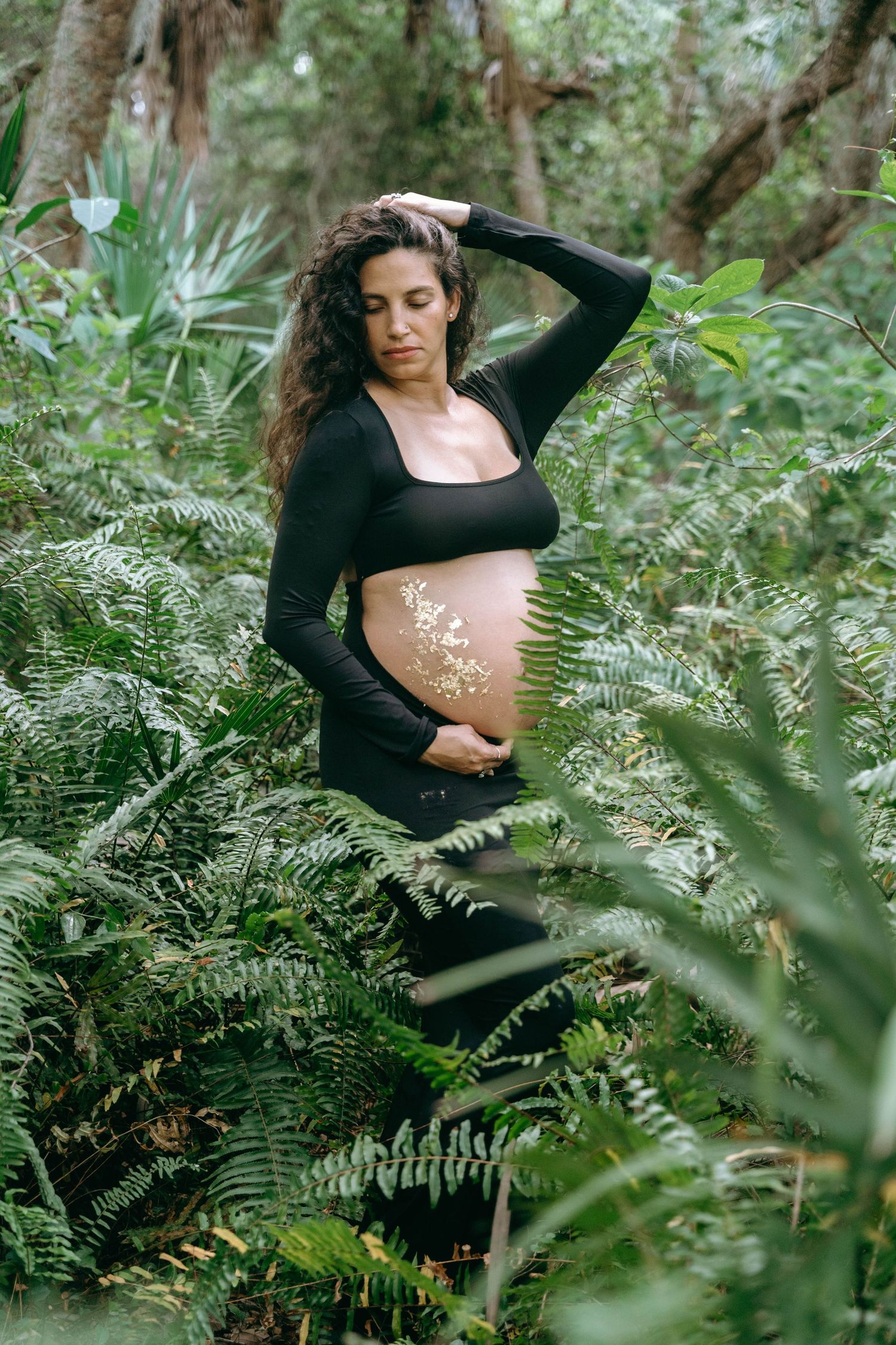 Woman in black outfit poses gracefully among lush green ferns in a forest setting.