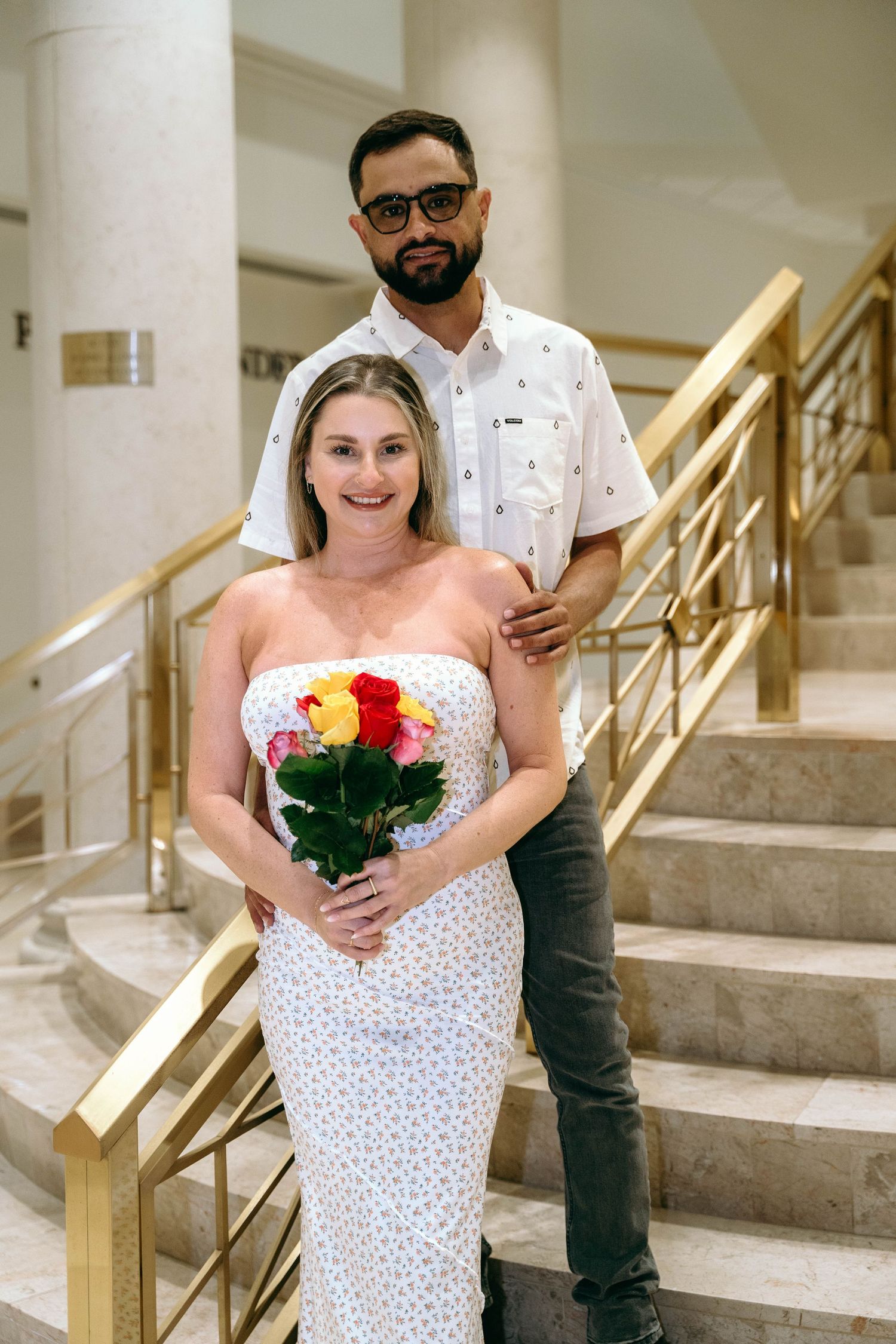 Bride in white polka dot dress holding colorful bouquet on marble staircase.