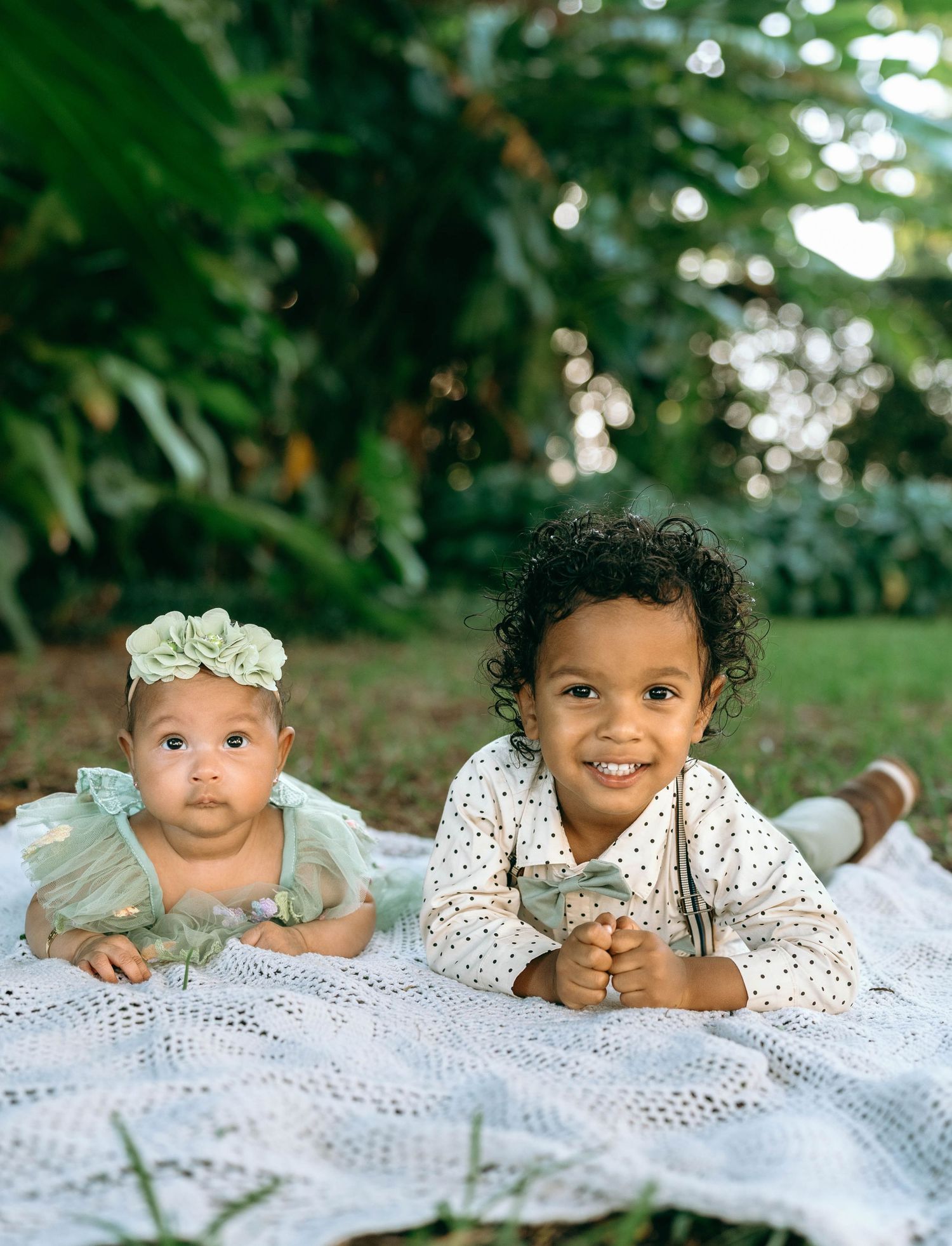 Two children lying on a white lace blanket outdoors with greenery and trees in the background.
