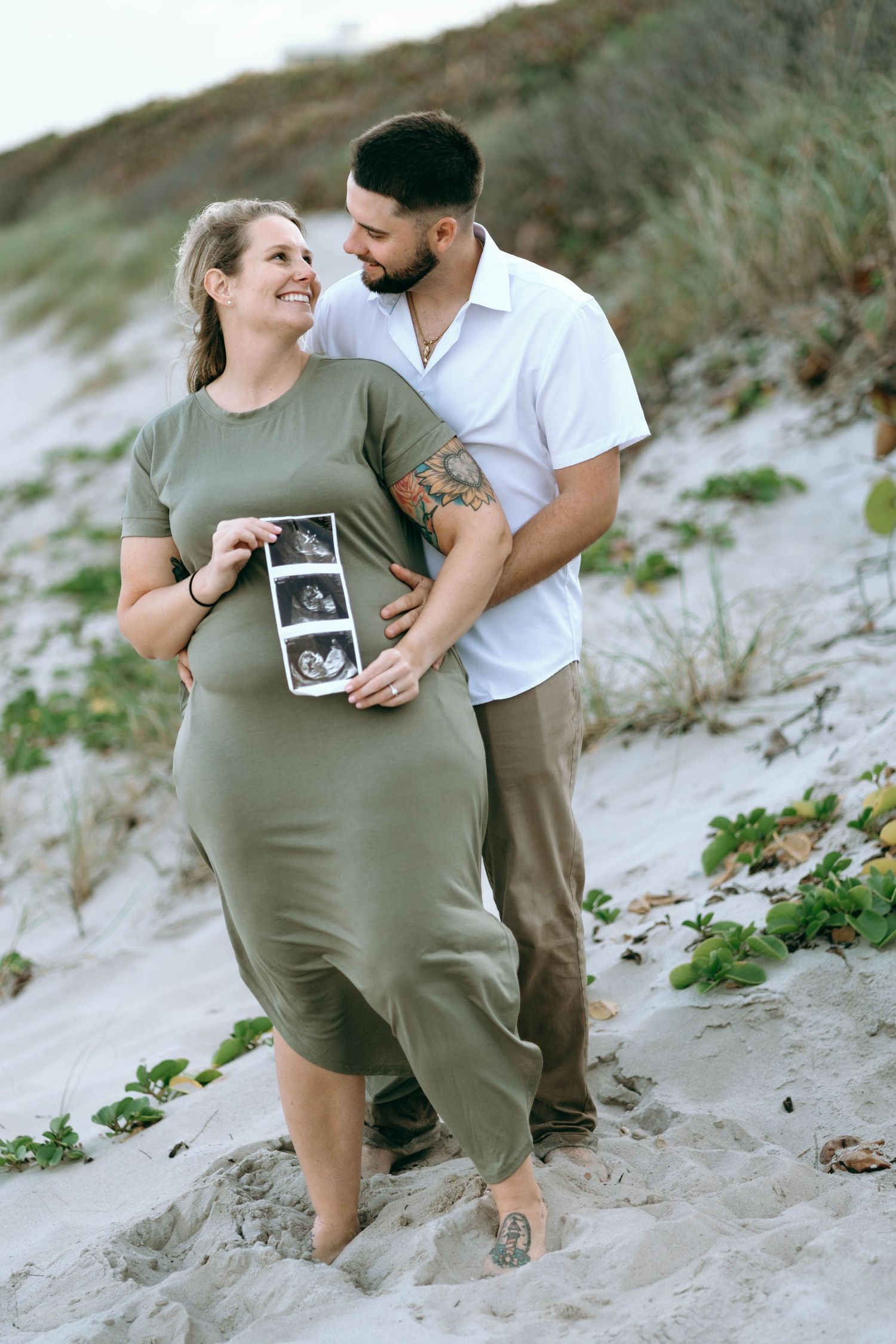 An expectant couple poses together on beach sand while holding ultrasound photos.