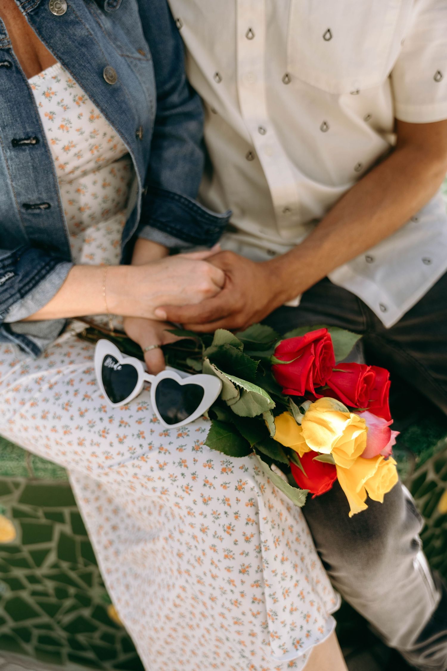Heart-shaped sunglasses and colorful roses rest on a floral dress with hands clasped together.