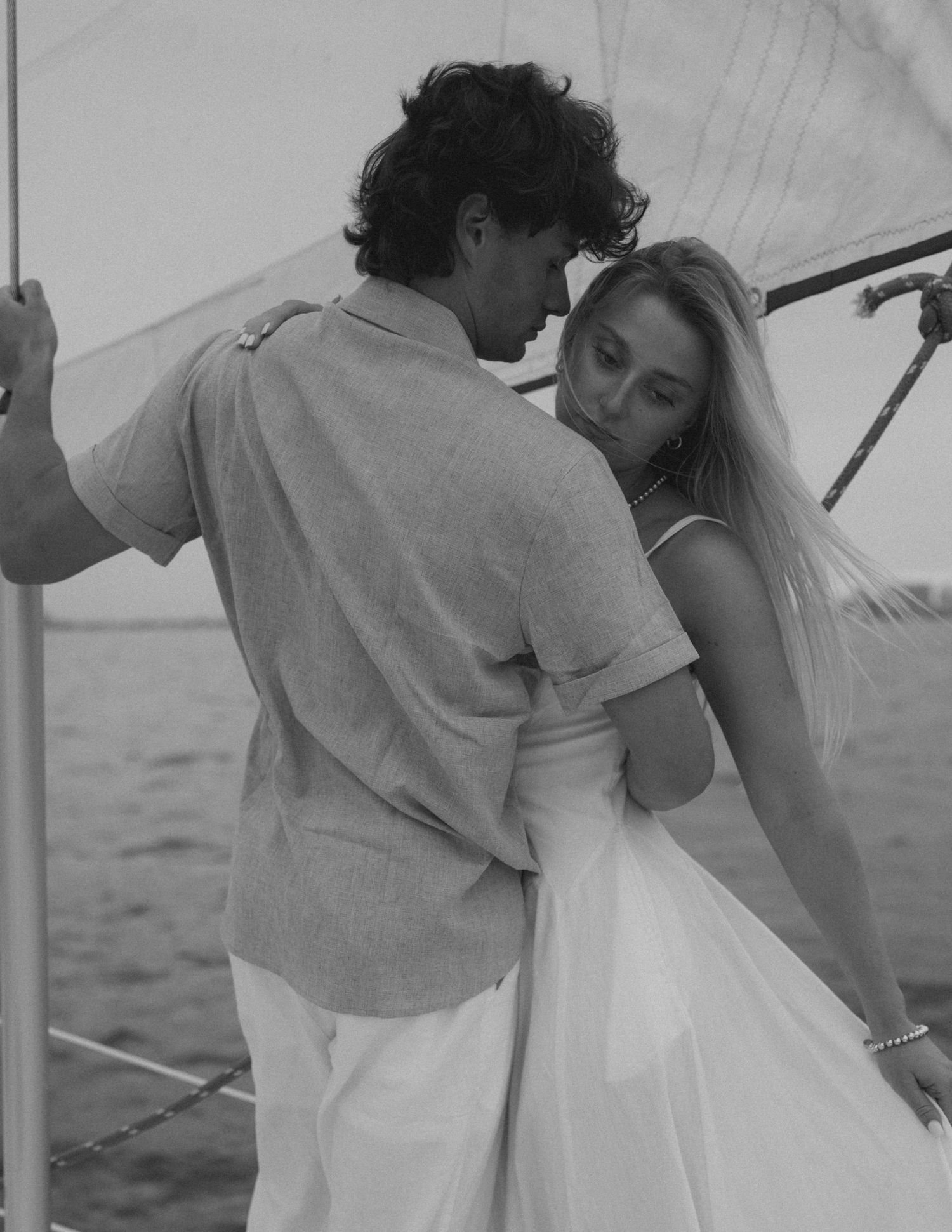 Black and white photo of a couple embracing romantically on a sailboat, woman in white dress, man in linen shirt.