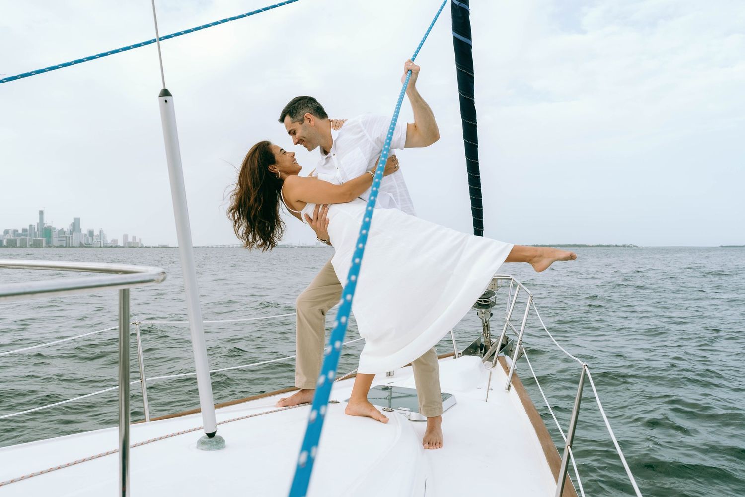 Couple dancing romantically on a white sailboat deck with ocean and city skyline in the background.