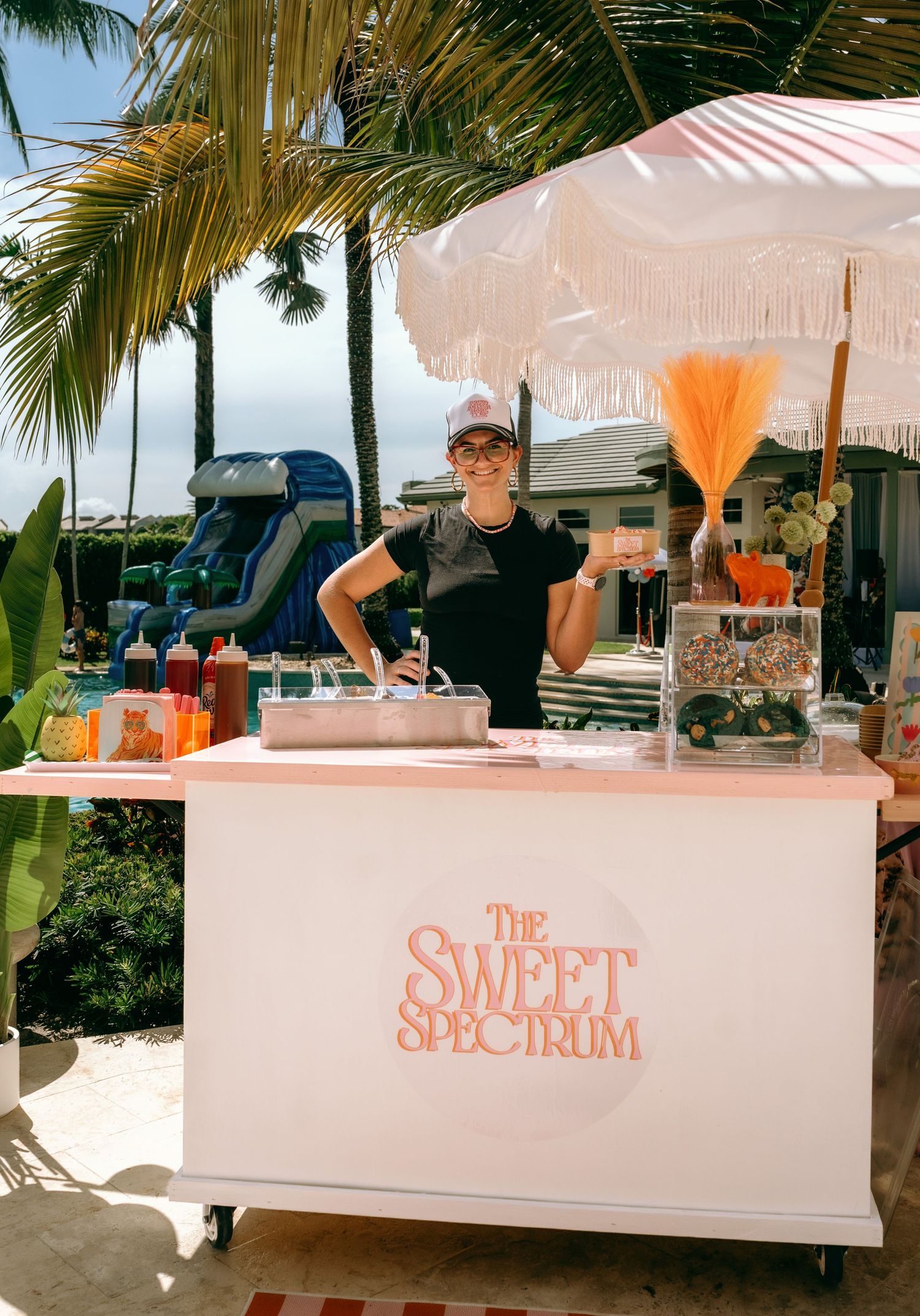 Woman smiling behind The Sweet Spectrum dessert cart under fringe umbrella at outdoor tropical event.