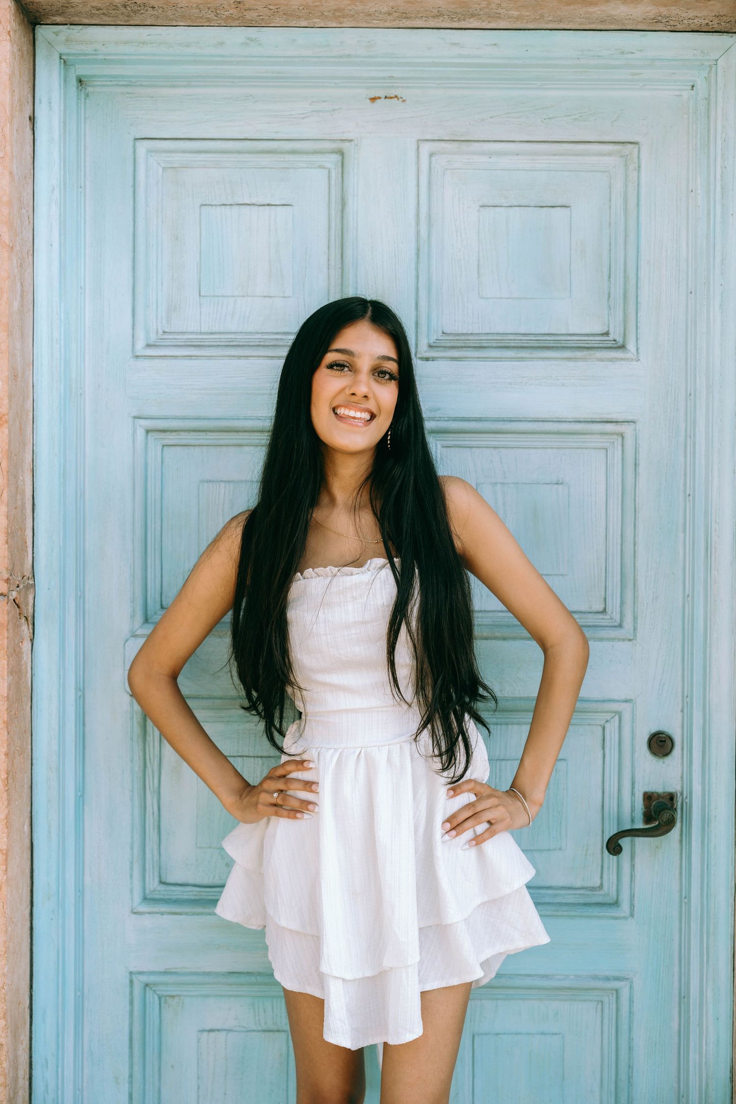 Smiling young woman with long dark hair in white strapless dress posing in front of a blue wooden door on Worth Avenue Palm beach.