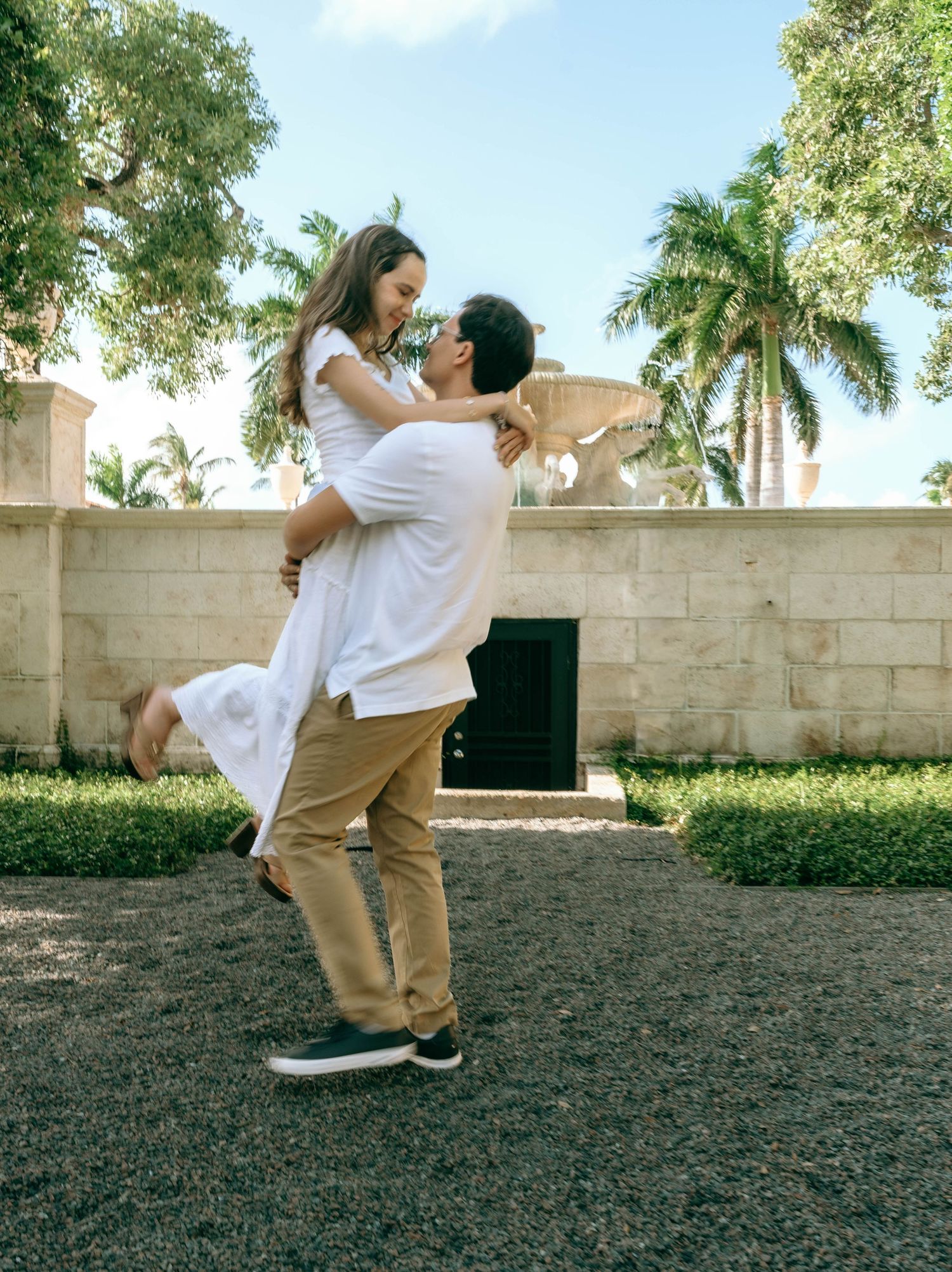 A romantic couple shares a joyful moment as a man lifts a woman in a white dress outdoors near palm trees. Engagement photos on Worth Avenue