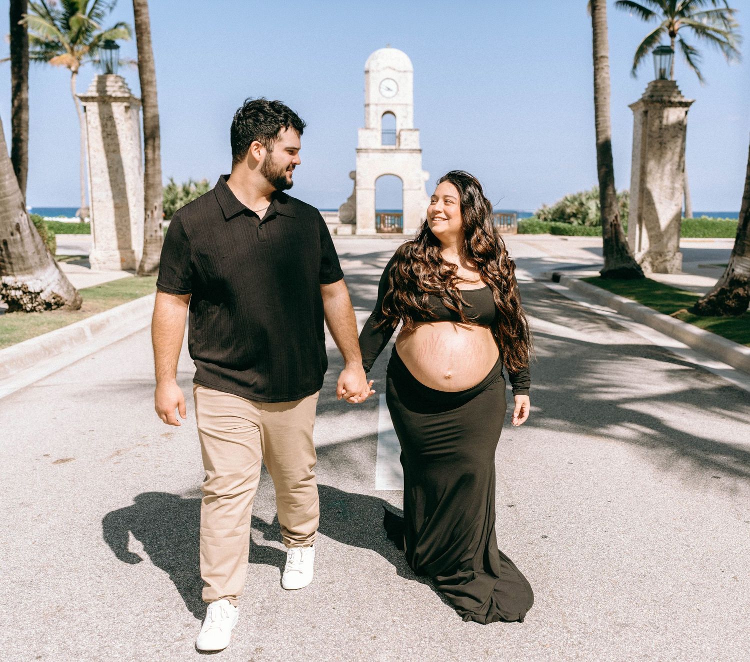 Pregnant couple holding hands walking outdoors near a clock tower with palm trees on a sunny day. Worth Avenue, Palm Beach, FL