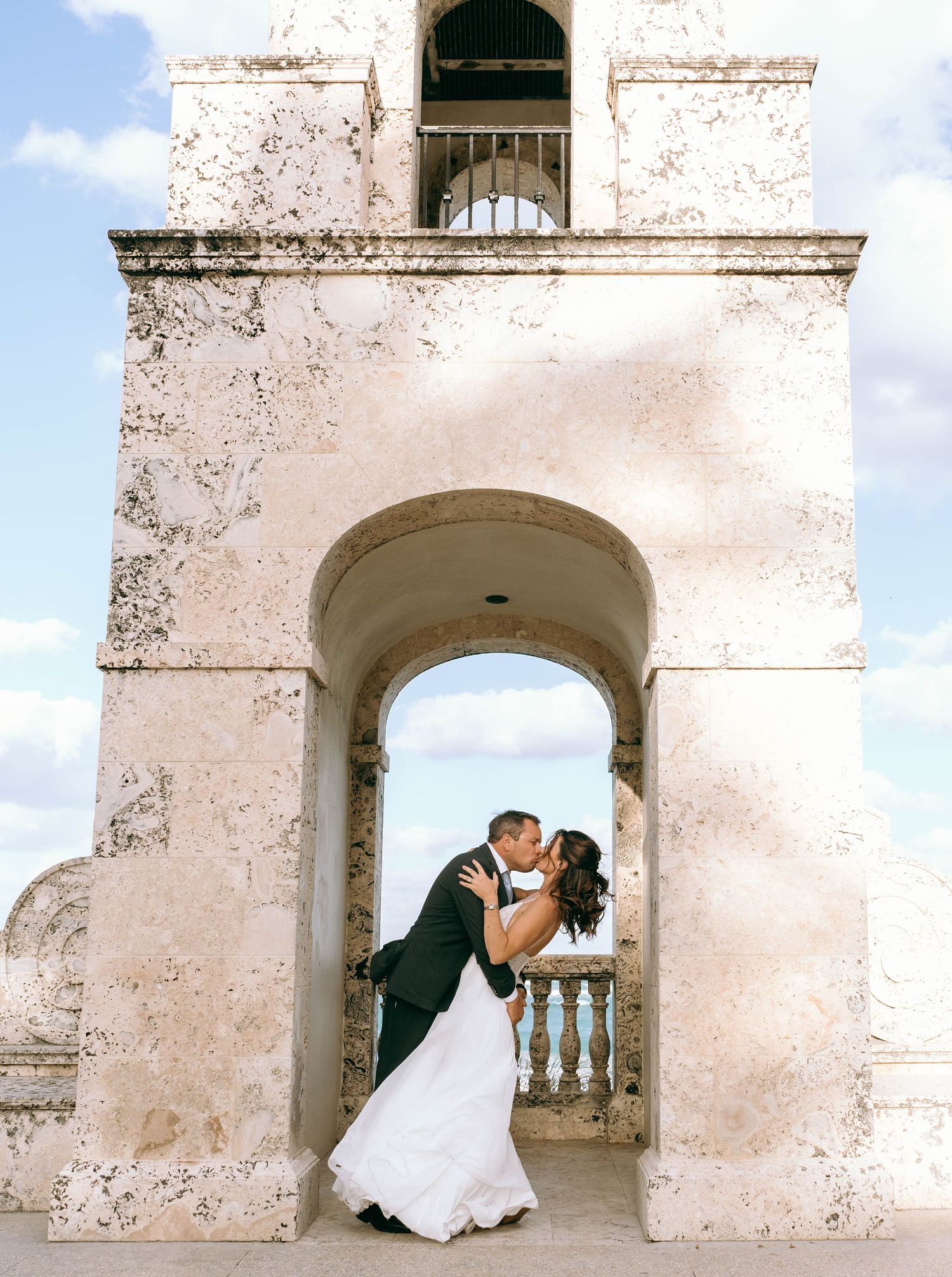 Bride and groom share a kiss under a historic stone archway with a bell tower and blue sky backdrop.