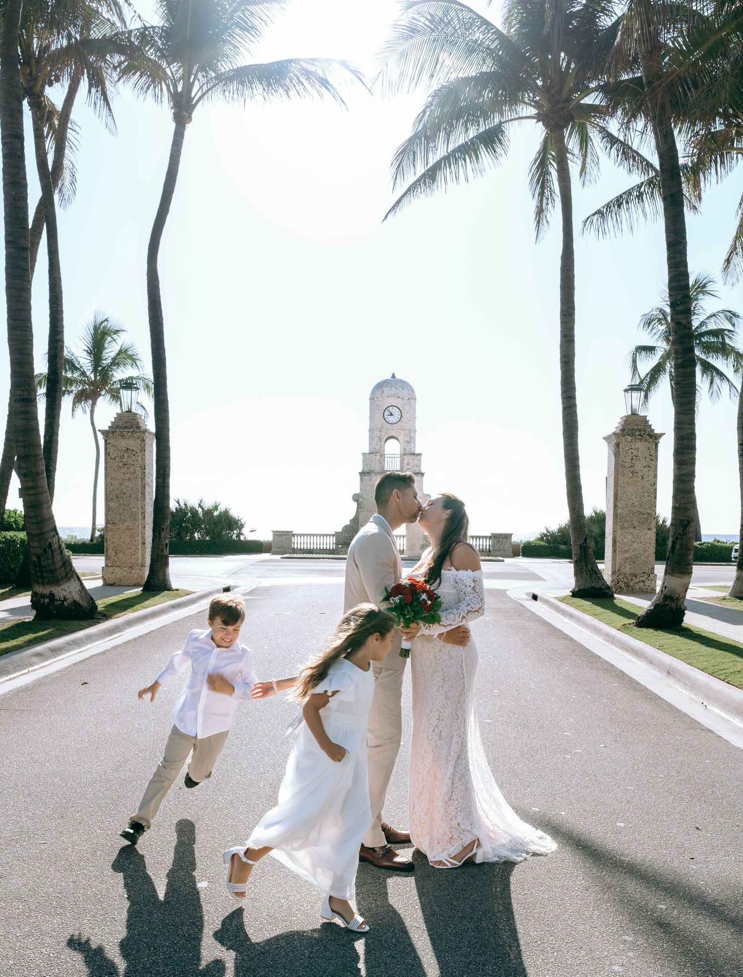Wedding couple kisses on palm-lined avenue as two children play around them near a clock tower.
