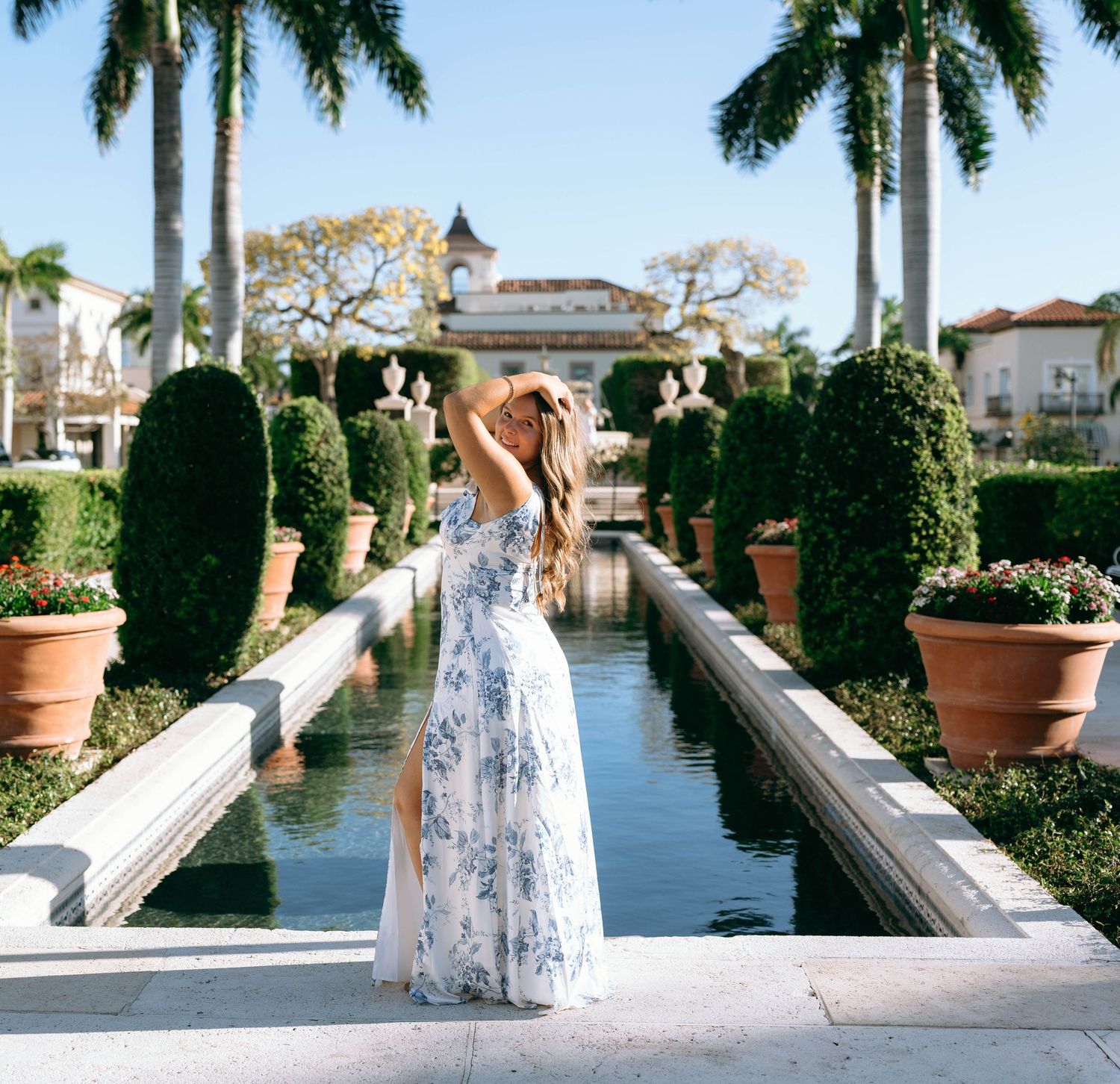 Woman in blue floral maxi dress poses by reflecting pool surrounded by palms and topiaries.