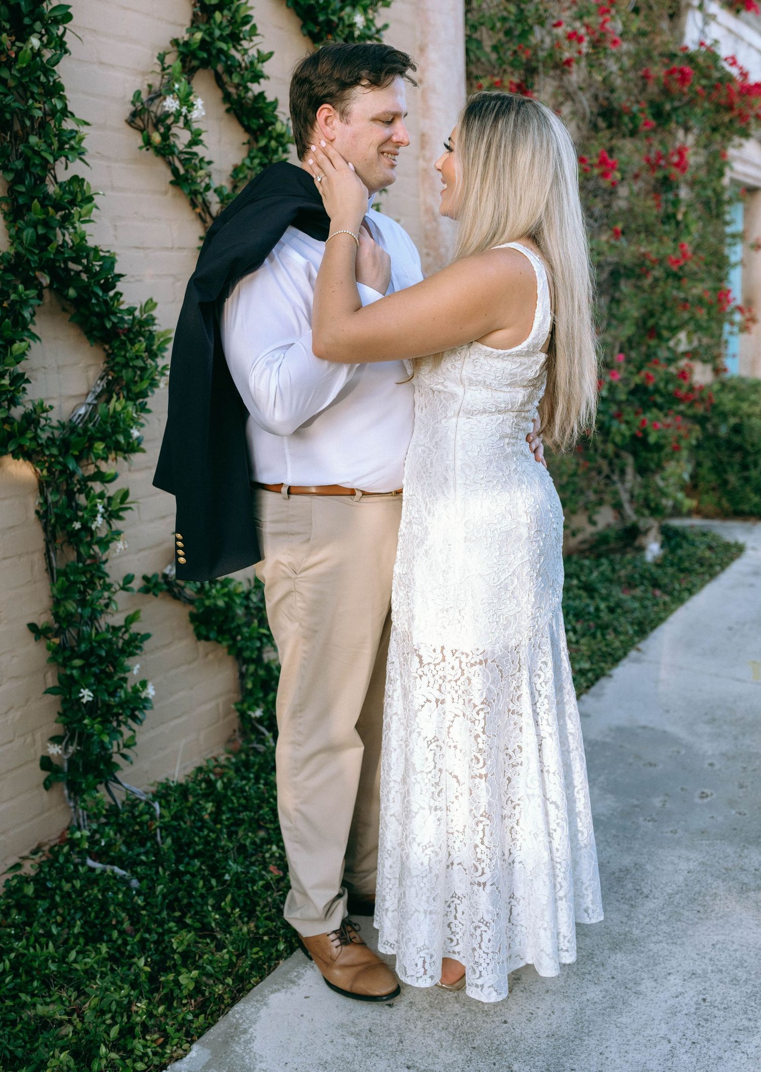 Couple sharing a romantic moment, woman in white lace dress touching mans face, ivy wall backdrop.