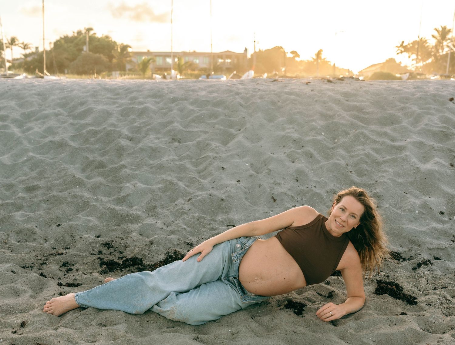 Pregnant woman in brown top and jeans posing on sandy beach at golden hour sunset.