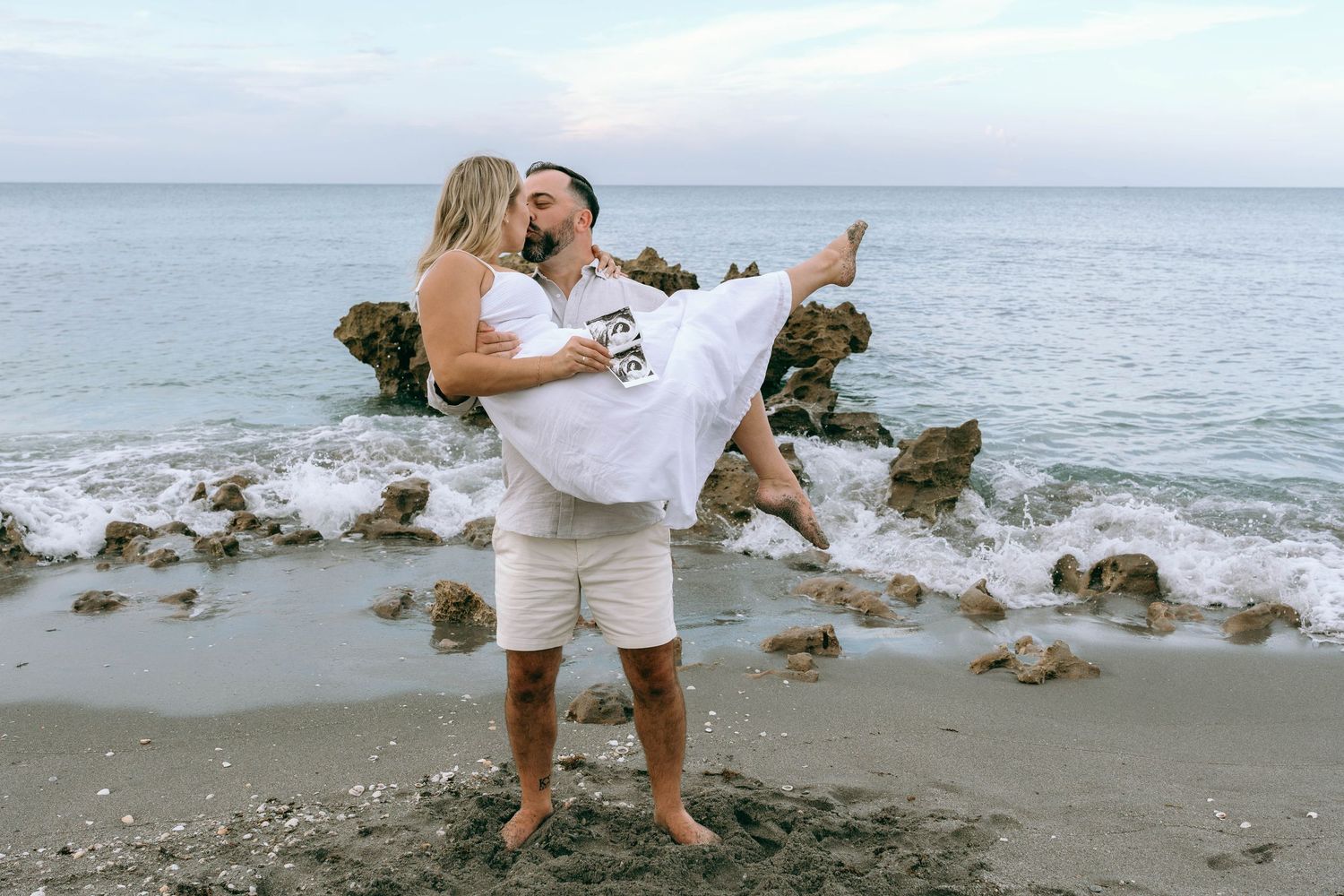 Romantic couple embracing on a rocky beach, man lifting woman in white dress near ocean waves.