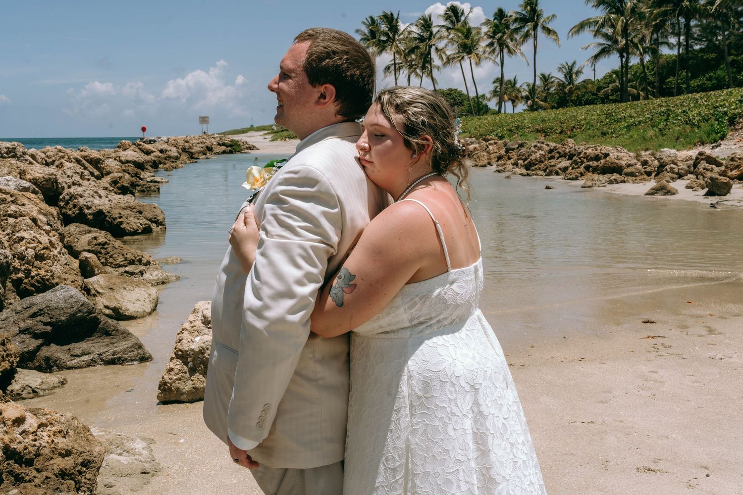 Bride and groom embracing on a rocky tropical beach with palm trees and blue ocean water behind them.