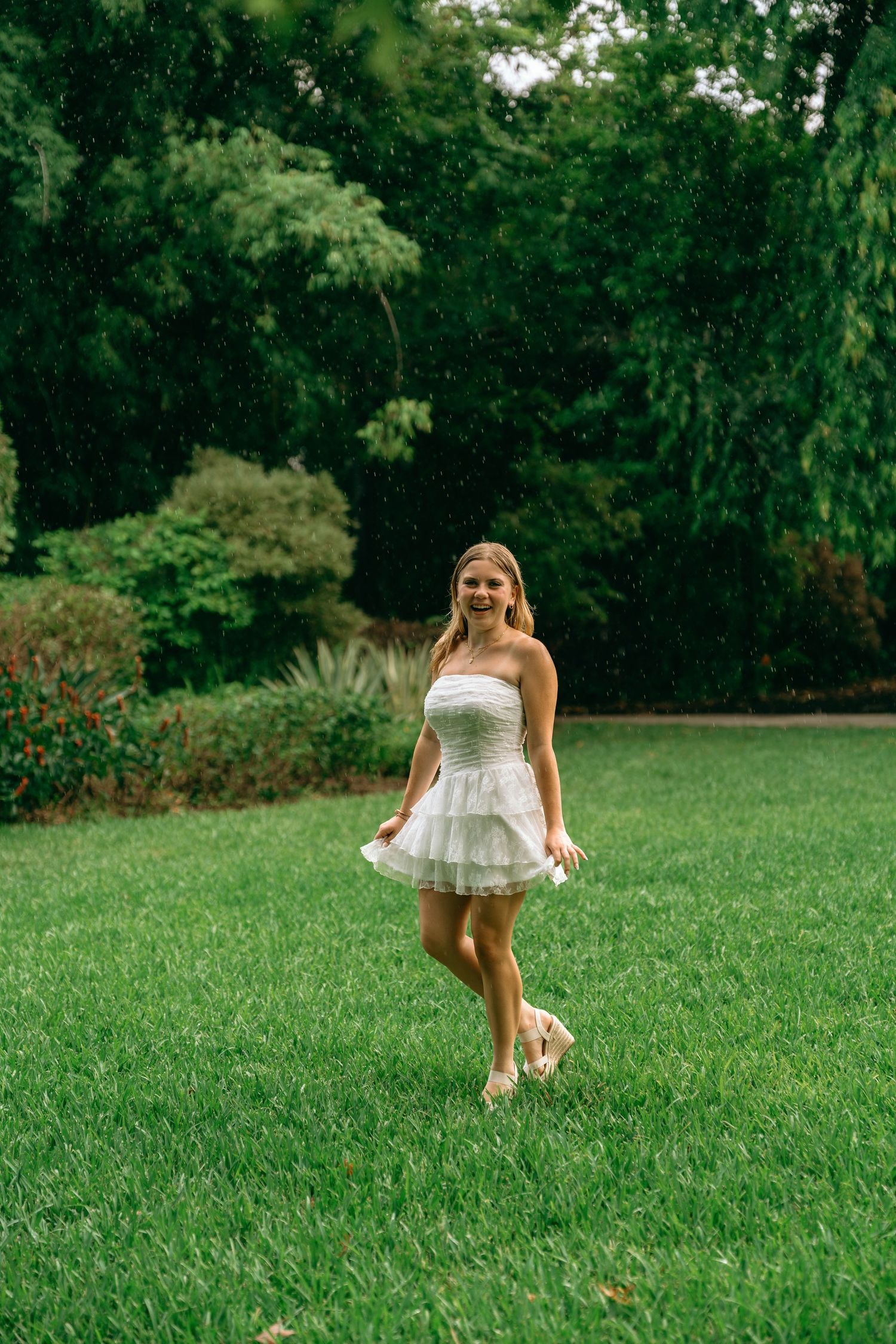 Young woman in white strapless mini dress twirling on lush green lawn surrounded by trees.