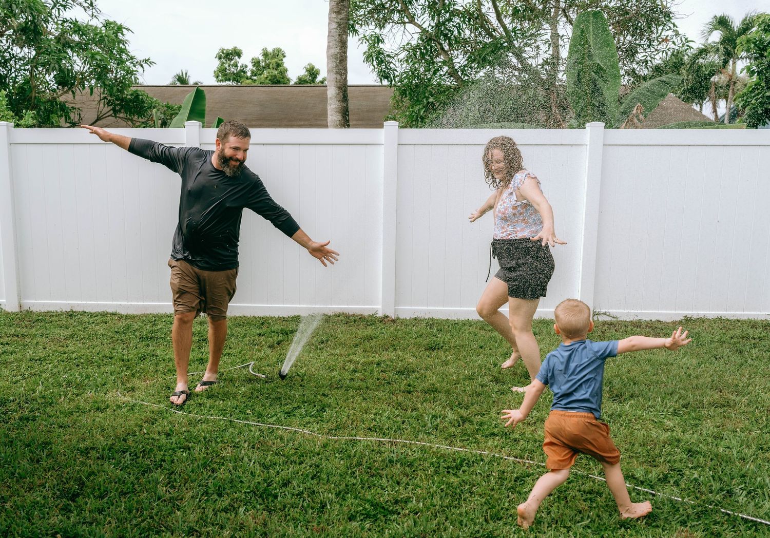Family playing with a sprinkler in a sunny backyard, with parents and toddler laughing and running on green grass.