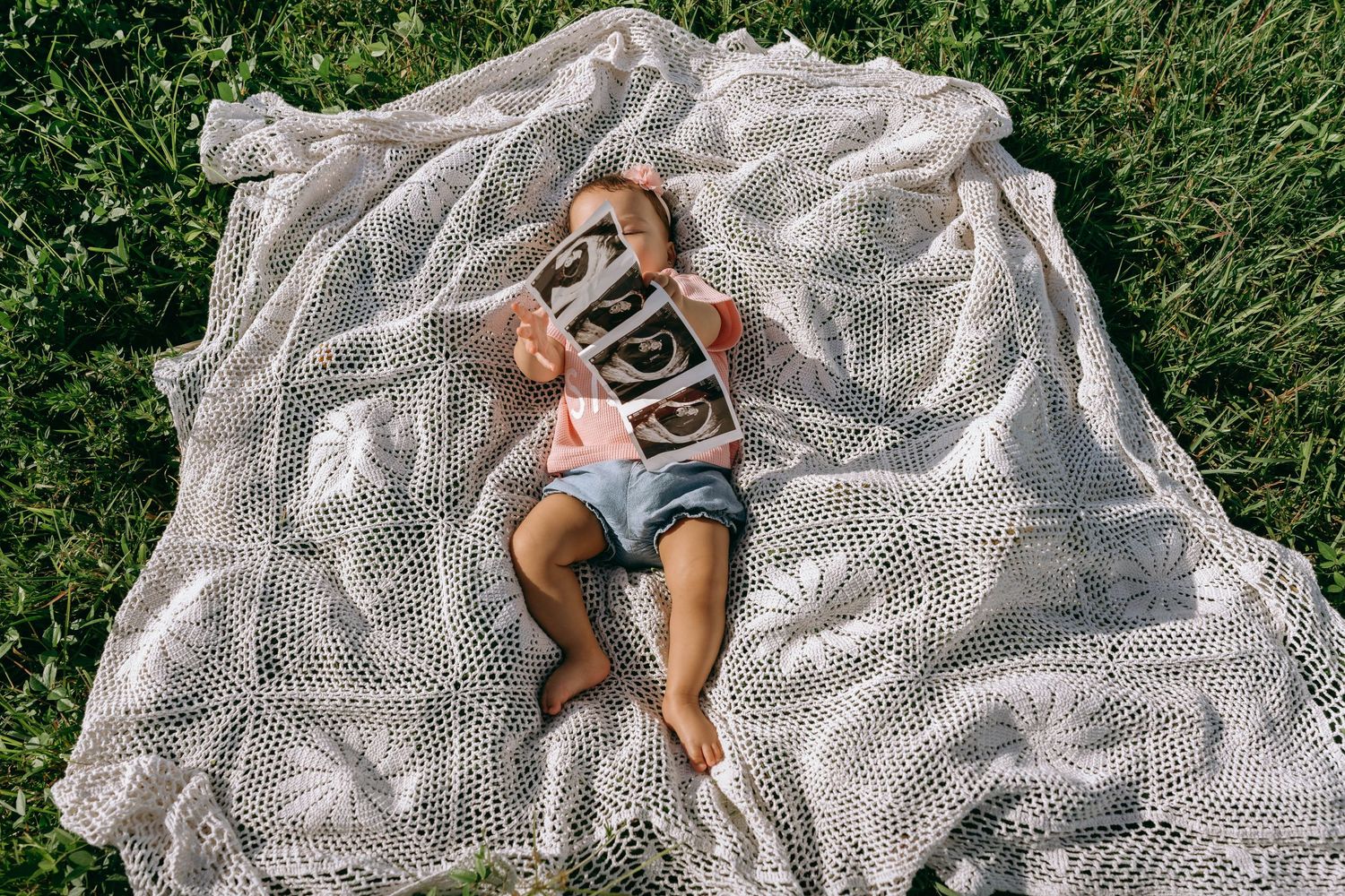 Baby lying on a white lace blanket in green grass, holding letter blocks spelling a name outdoors.