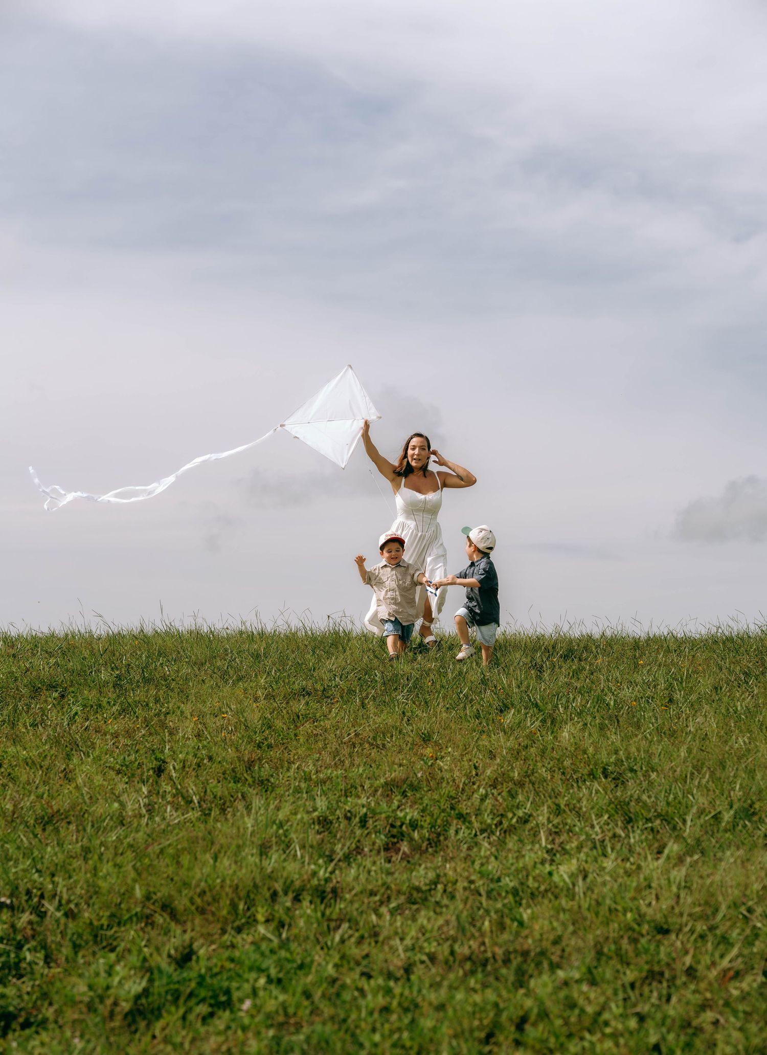 A woman in a white dress flies a white kite with two young children on a green grassy hill under a cloudy sky.