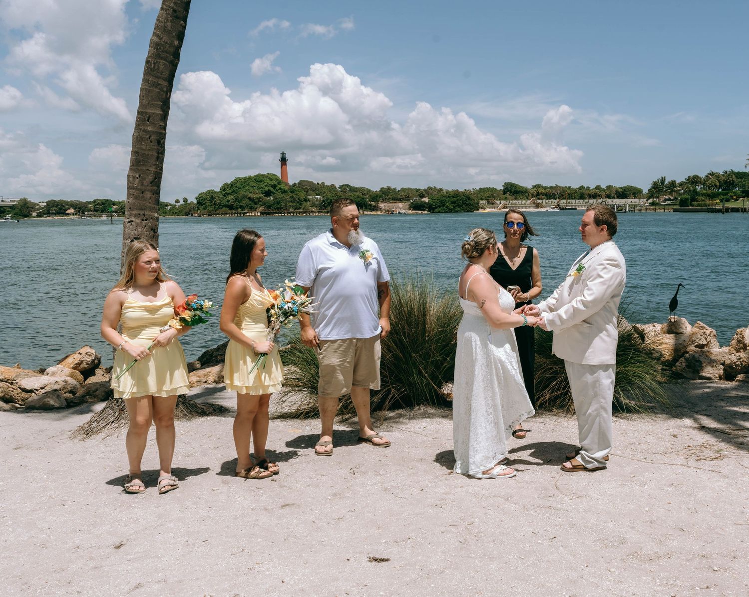 Outdoor waterfront wedding ceremony with bride, groom, officiant, and bridesmaids in yellow dresses near a lighthouse.