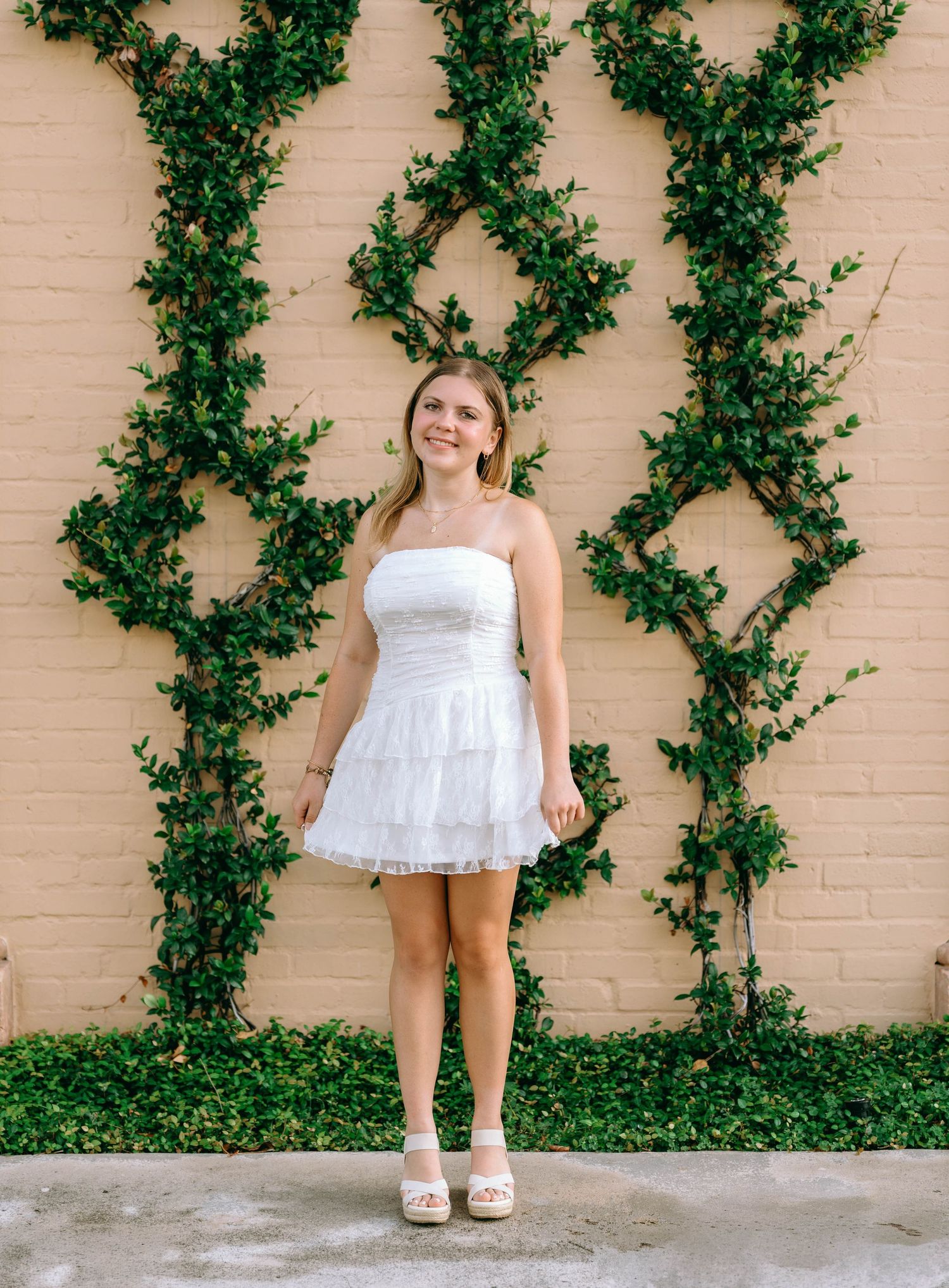 Young woman in white strapless tiered mini dress posing against brick wall with green ivy vines.
