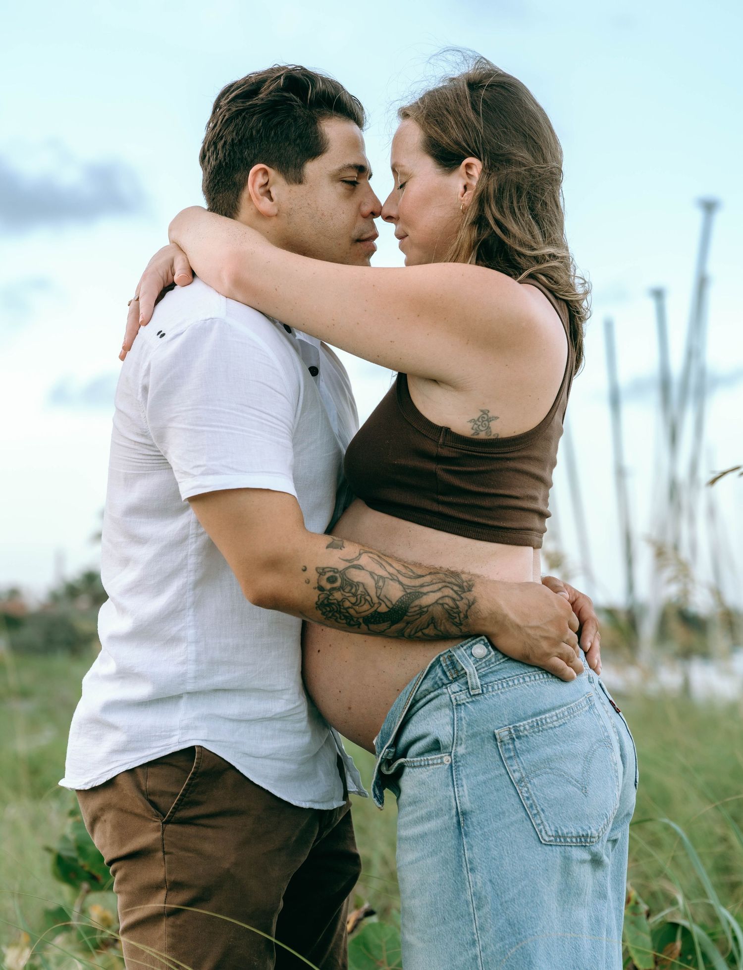 Pregnant couple embracing outdoors, woman with tattoos in crop top and jeans, man in white shirt, sharing a tender moment.