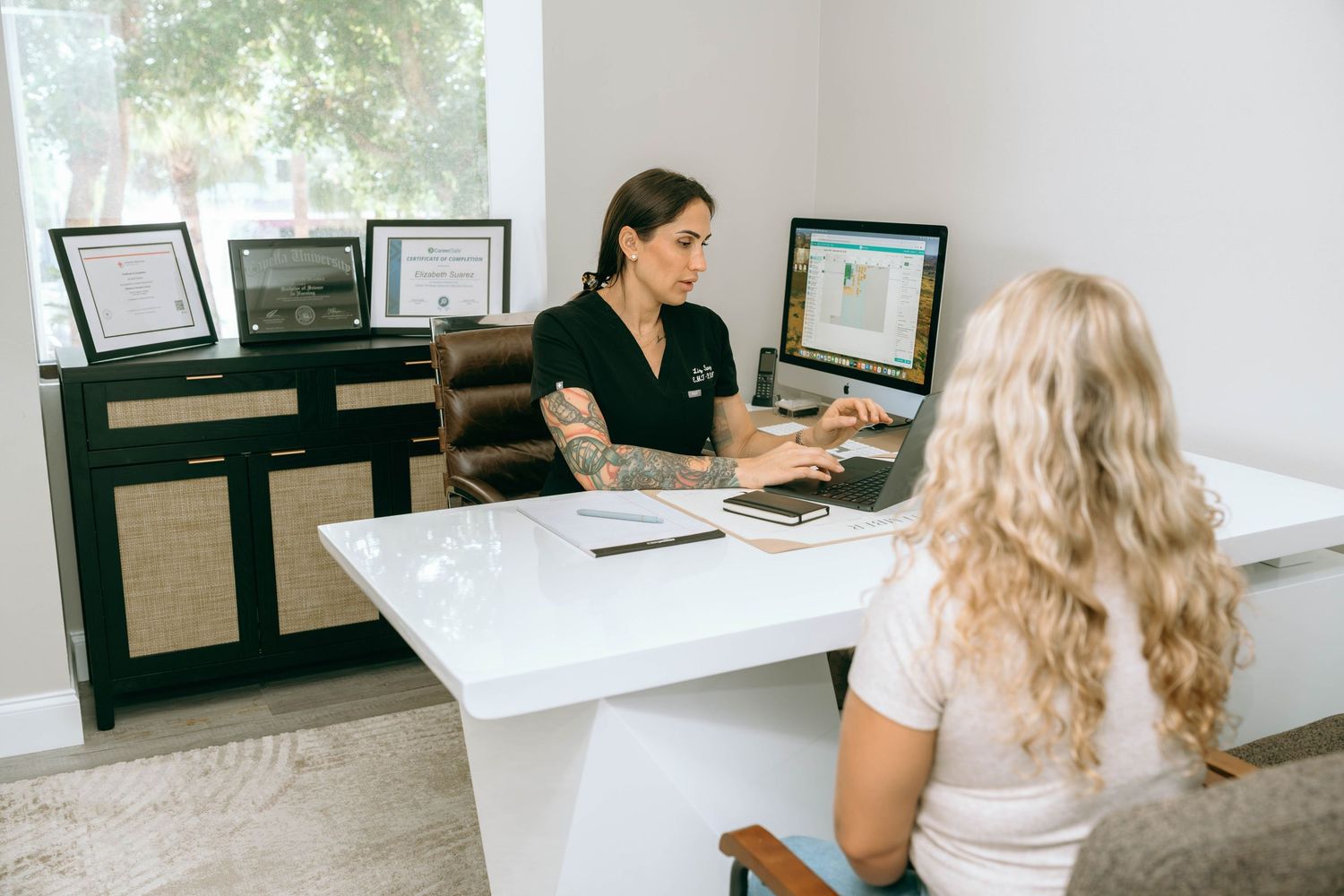 A tattooed receptionist consults with a blonde client at a modern white desk in a bright medical or wellness office.