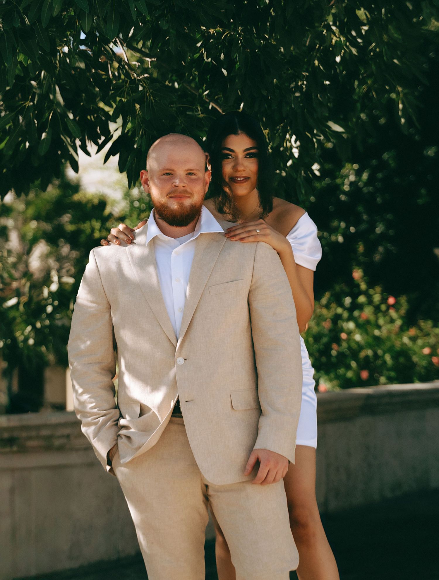 Couple in beige suit and white dress standing among lush greenery.