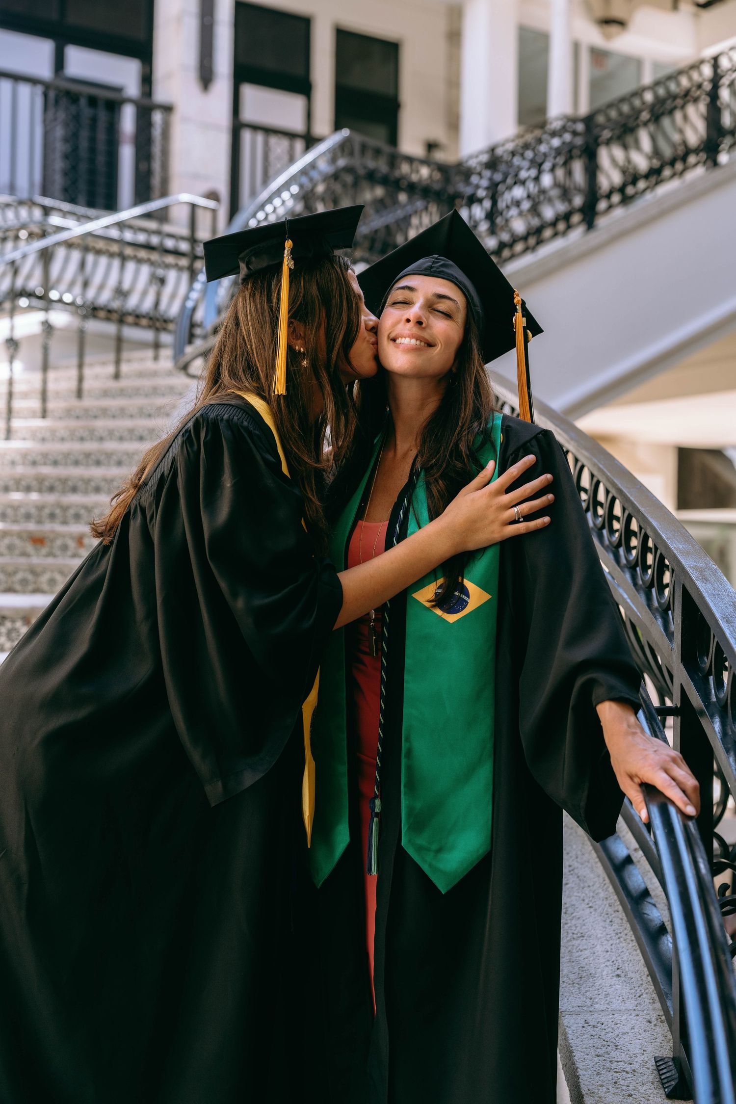 Two female graduates in black caps and gowns embracing on outdoor staircase on Worth Avenue Palm beach.