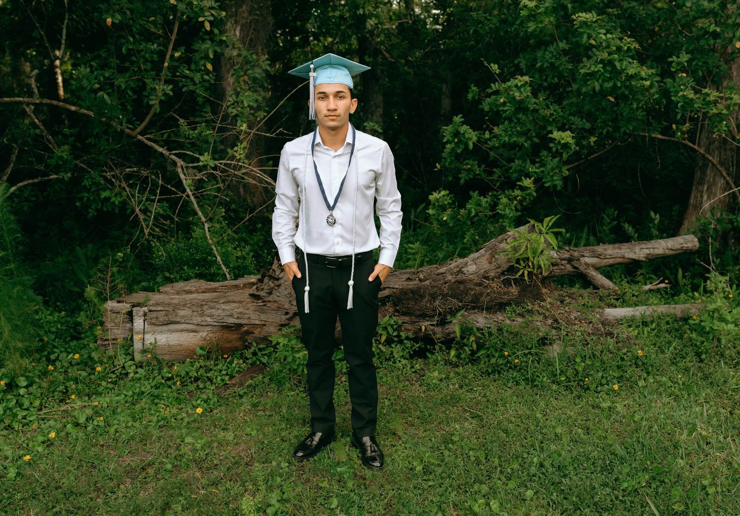 Young man in graduation cap and gown with medal stands proudly outdoors in a lush green forest setting. Senior Photos at Colt Farms