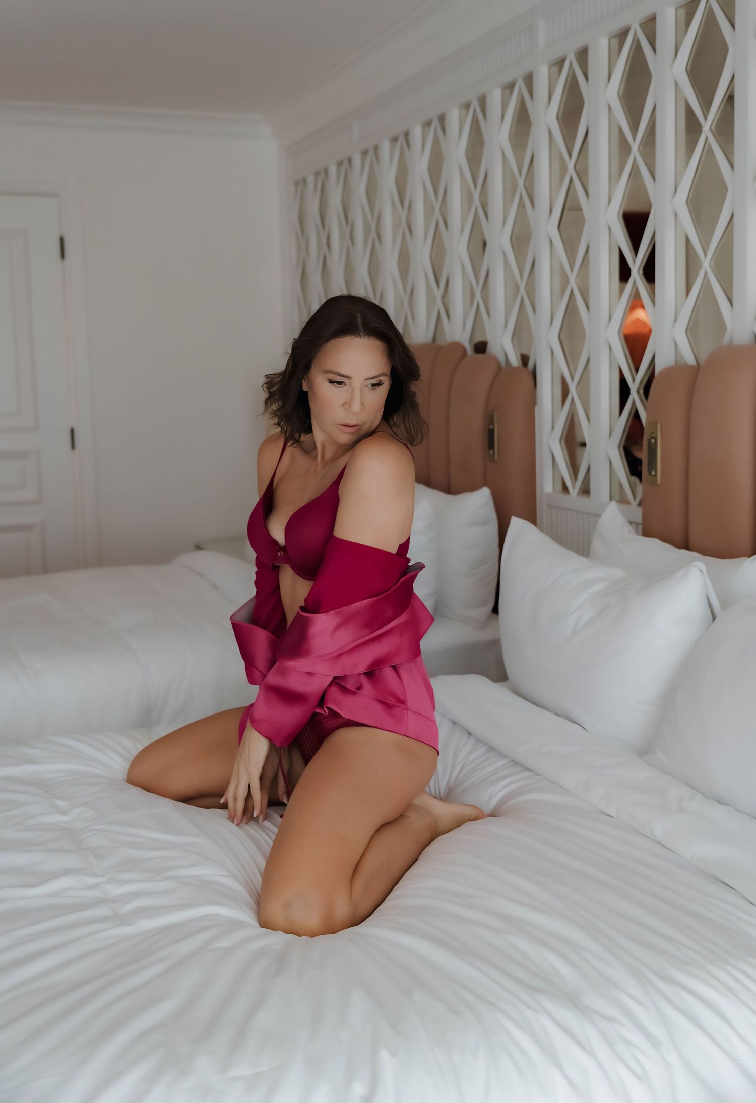 Woman in pink satin lingerie and robe posing on white bedding in an elegant hotel room.  Boudoir Palm Beach, FL