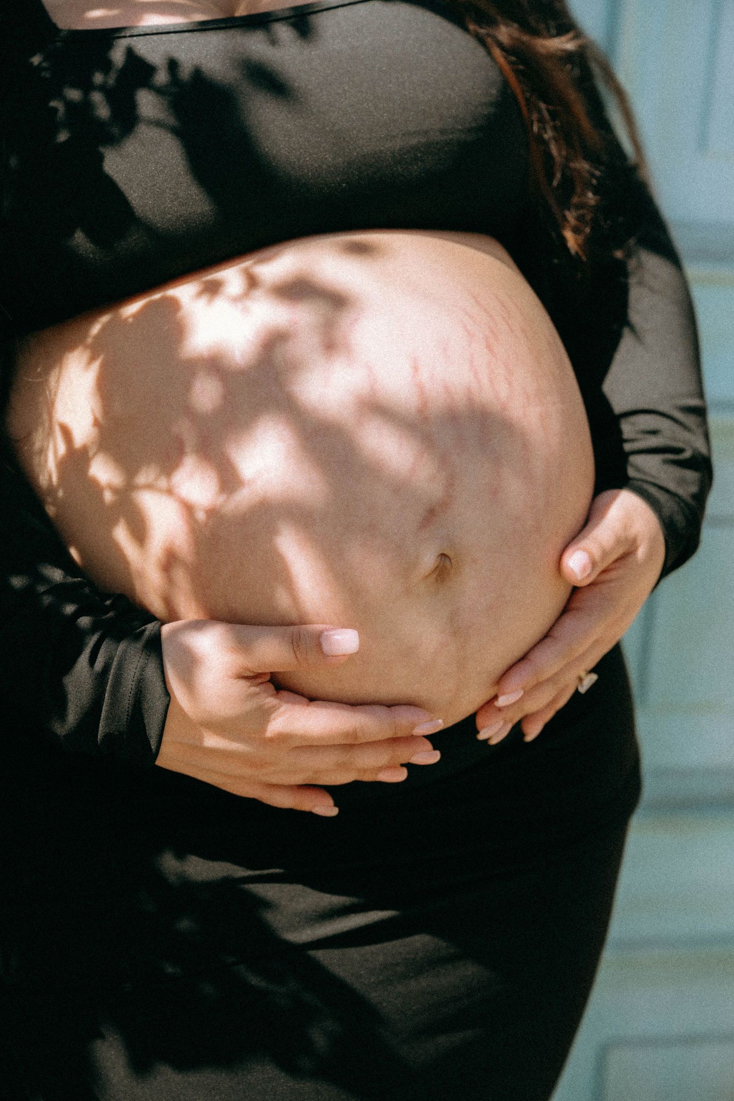 Pregnant woman in black outfit cradling her bare baby bump with dappled sunlight casting leaf shadows. Worth Avenue, Palm Beach, FL
