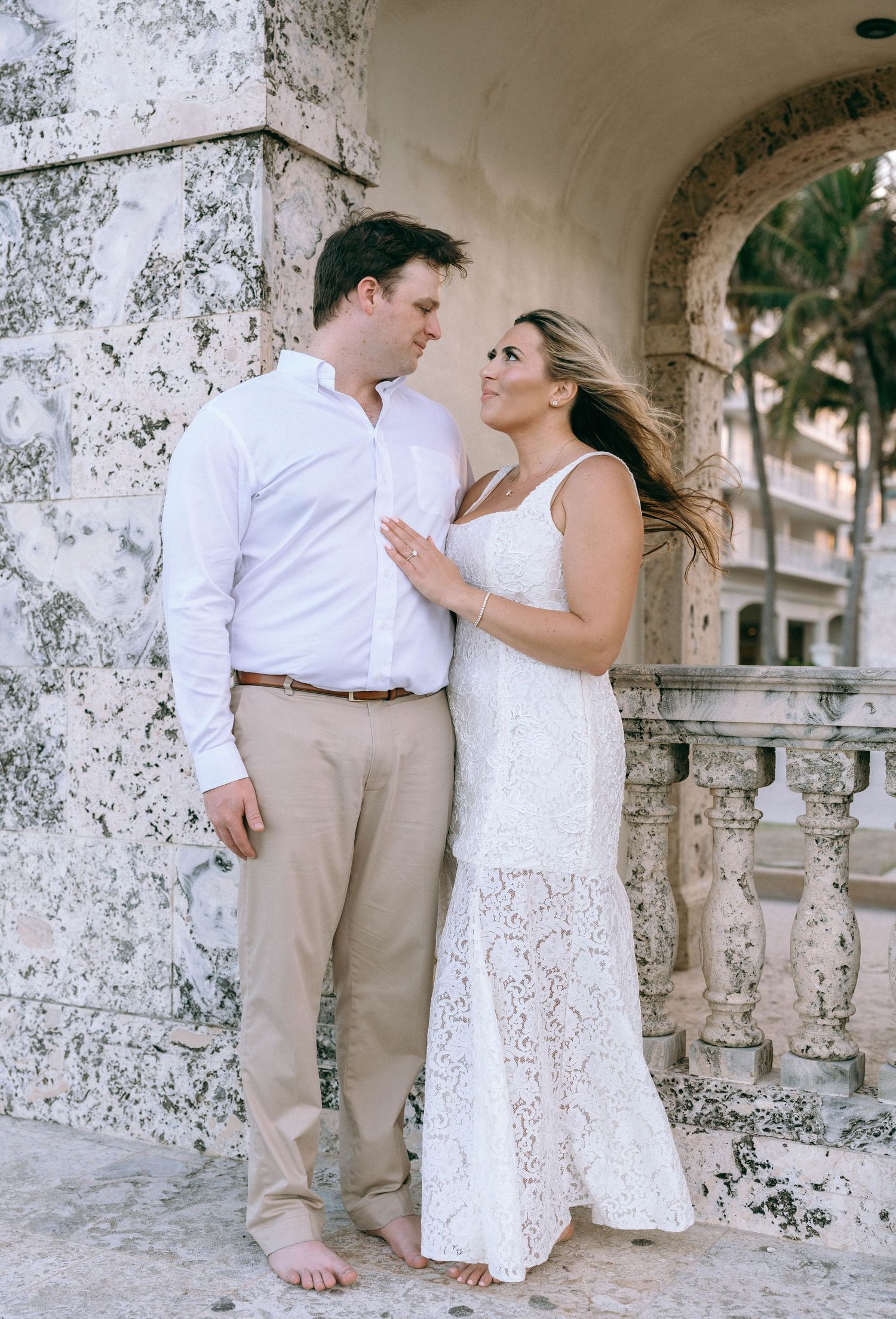 Couple gazing at each other by stone archway, woman in white lace dress, hair blowing in wind.