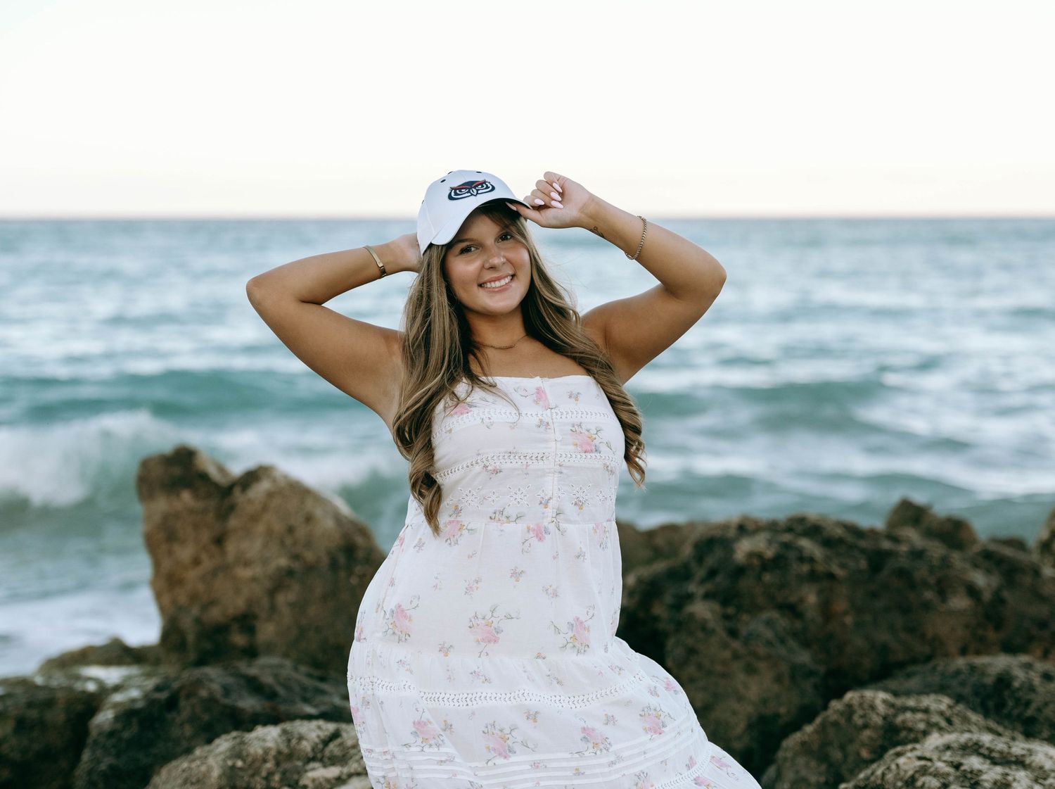 Woman in white floral dress and cap poses on rocky shoreline with ocean backdrop. Worth Avenue, Palm Beach, FL