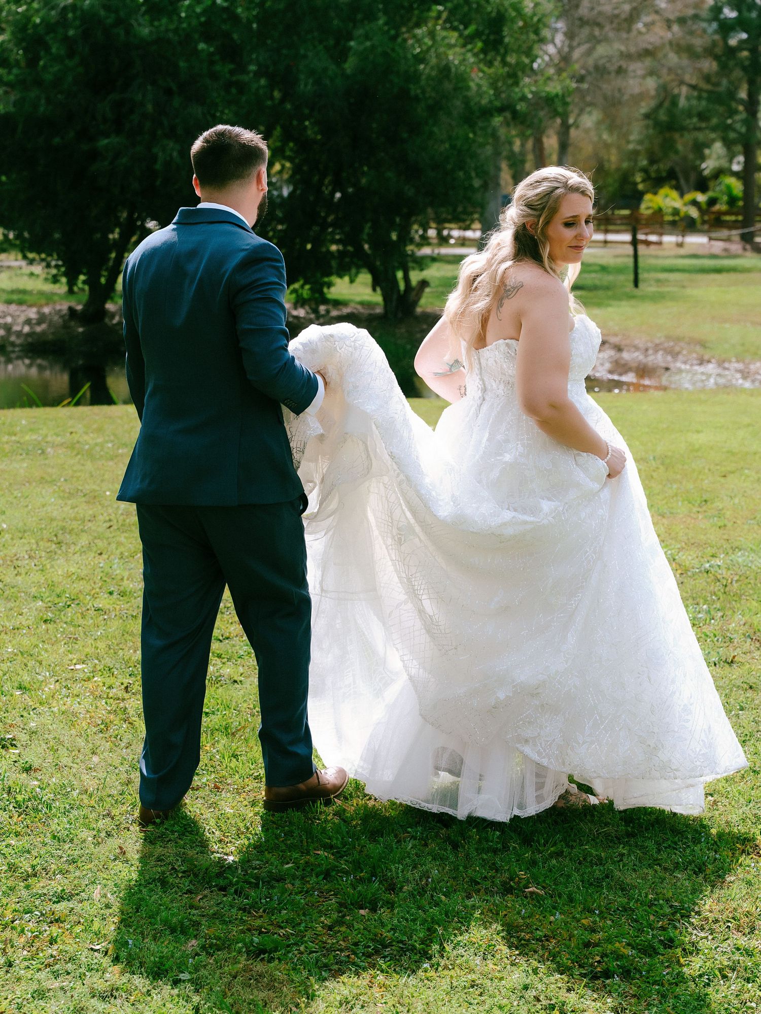 Groom in navy suit helps bride in lace ball gown walk across green lawn during outdoor wedding photos.