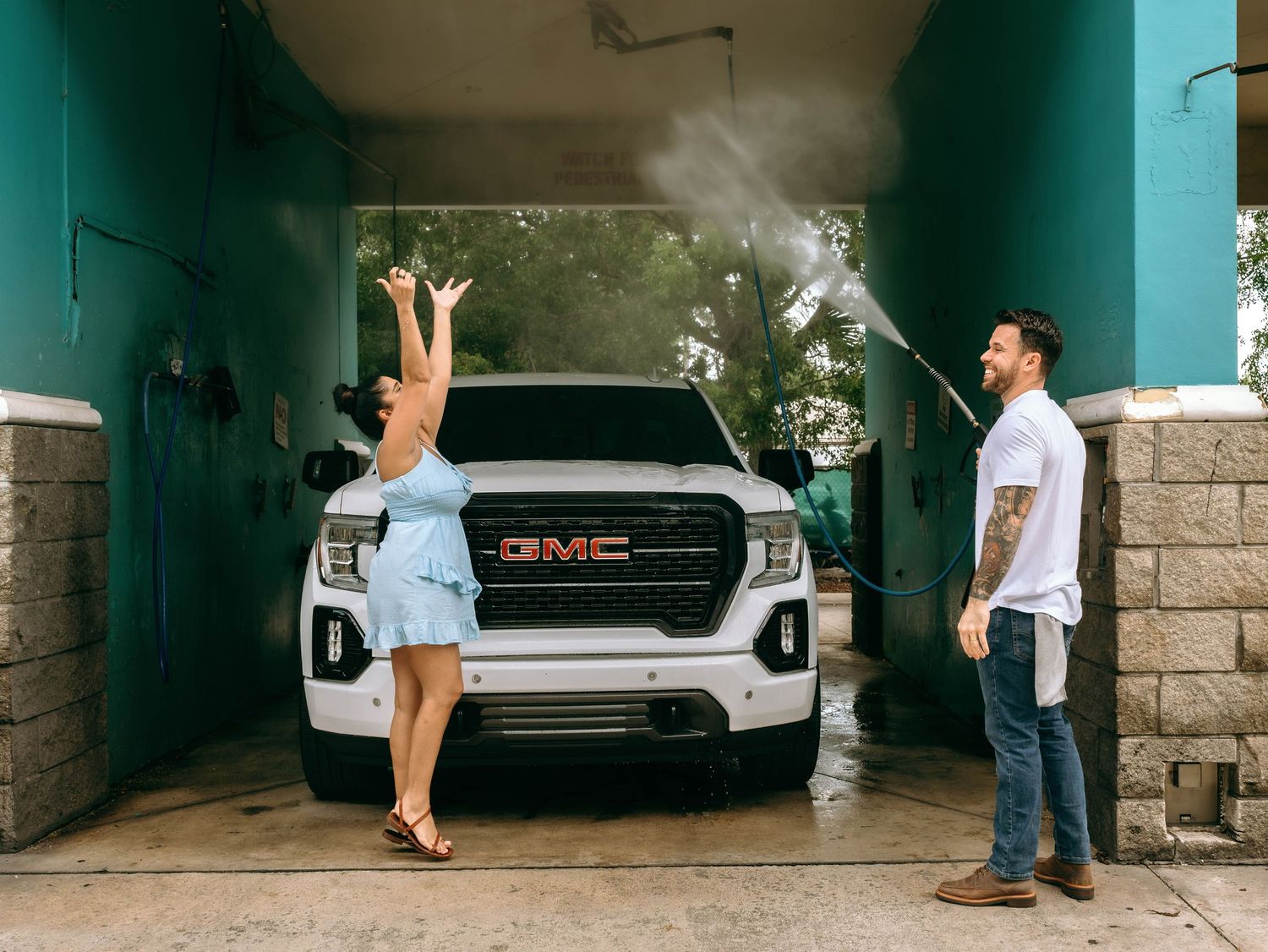 A woman in a blue dress poses with raised arms in front of a white GMC truck at a car wash with a man nearby.