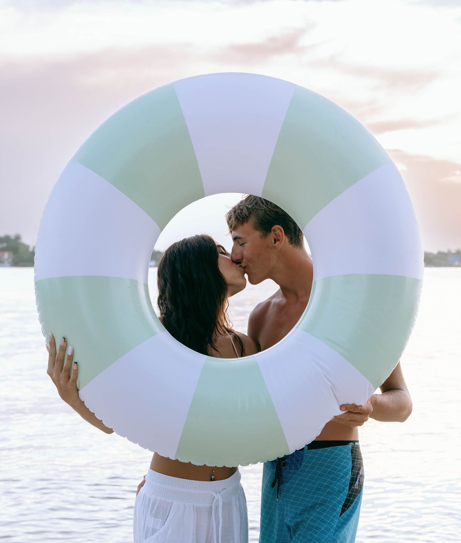 Couple kissing behind a mint striped inflatable pool ring near the ocean on a cloudy day.
