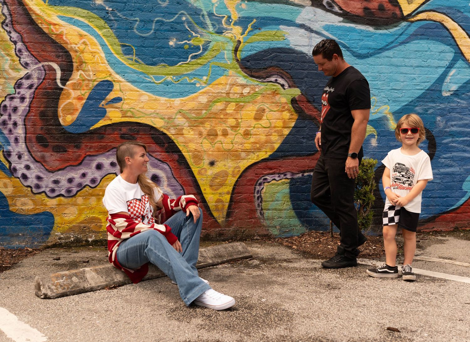 Two men and a young child pose in front of a colorful graffiti mural featuring an octopus on an urban wall.