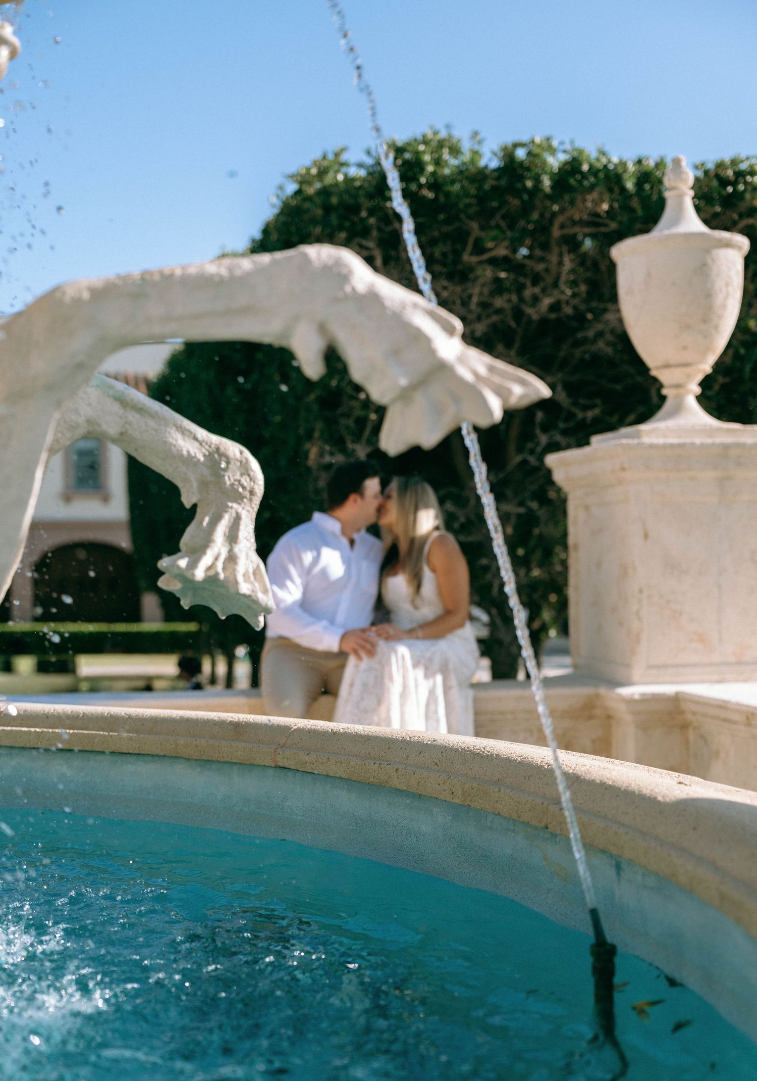 Couple sharing a romantic kiss behind an ornate stone fountain in a sunlit garden.