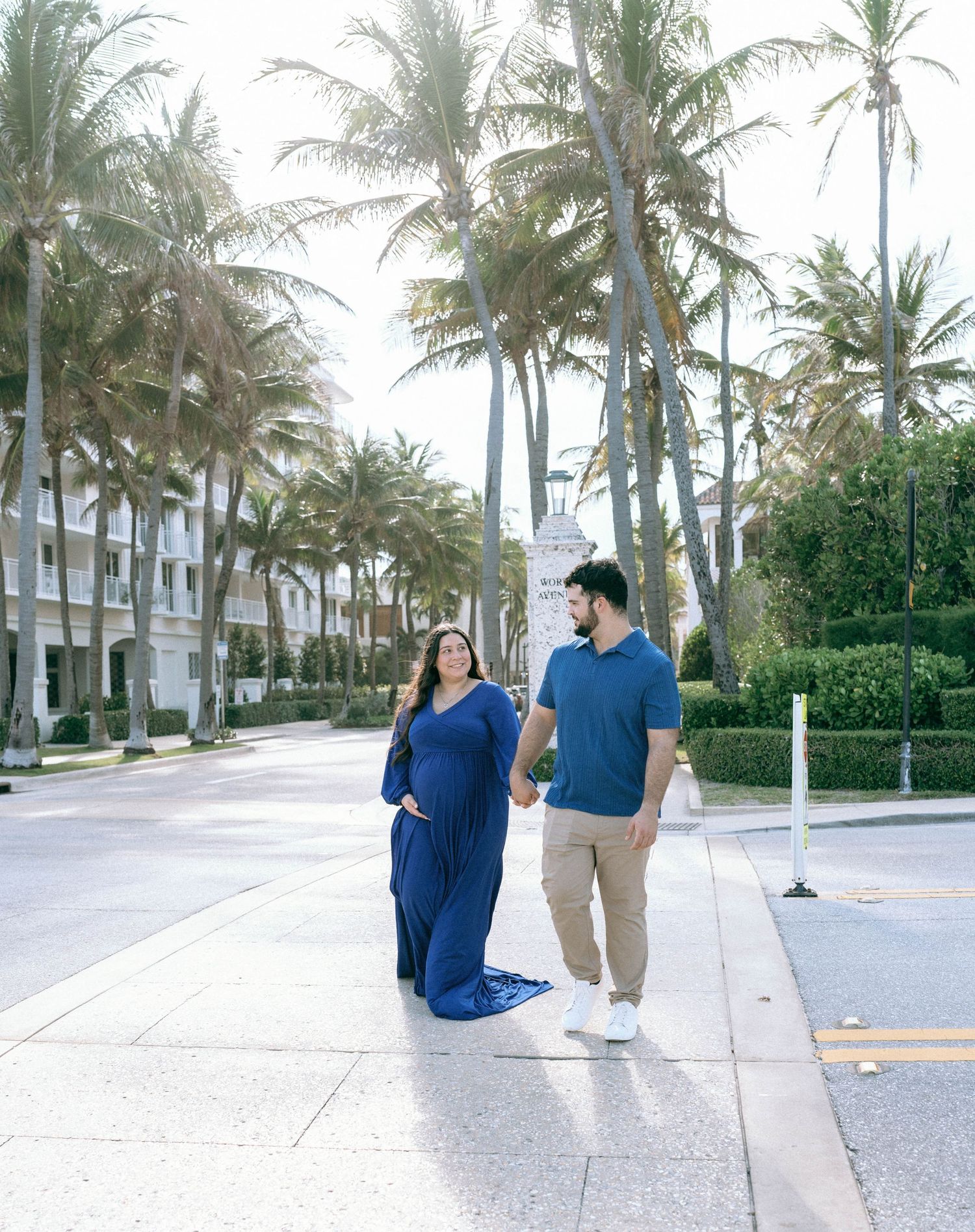 Expecting couple walking hand-in-hand along palm tree-lined street, woman in flowing blue maternity dress. Worth Avenue, Palm Beach, FL