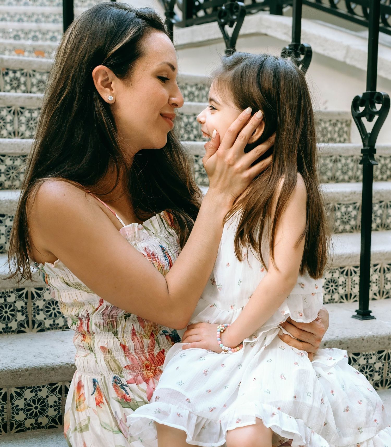 A mother in a floral dress tenderly holds her smiling daughter's face while sitting on decorative steps. Worth Avenue, Palm Beach, FL