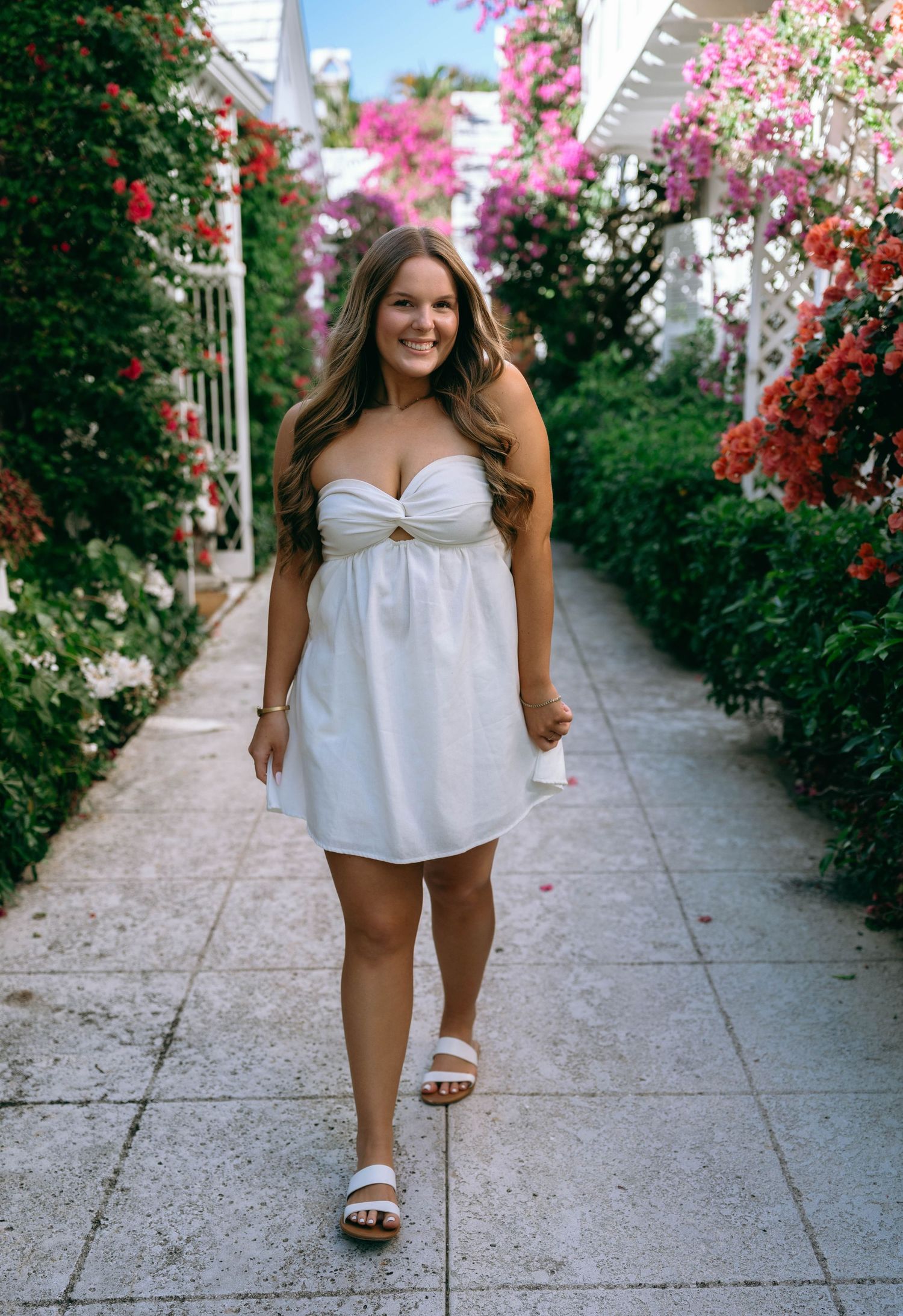 Smiling woman in white strapless mini dress posing on a flower-lined garden path.