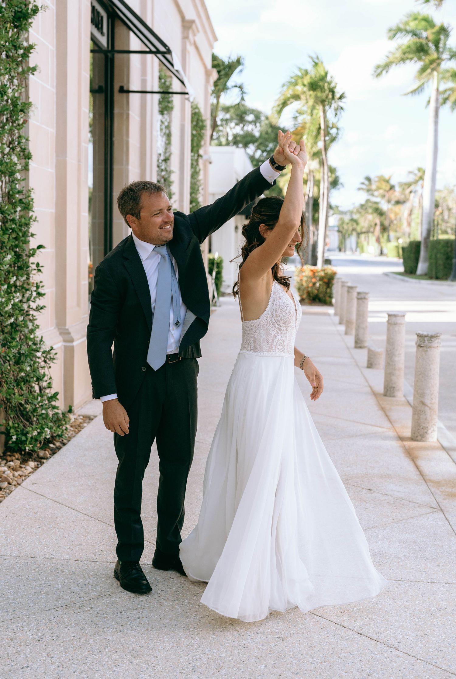 Groom twirls bride in white gown outside classical building with palm trees at sunny Florida wedding.